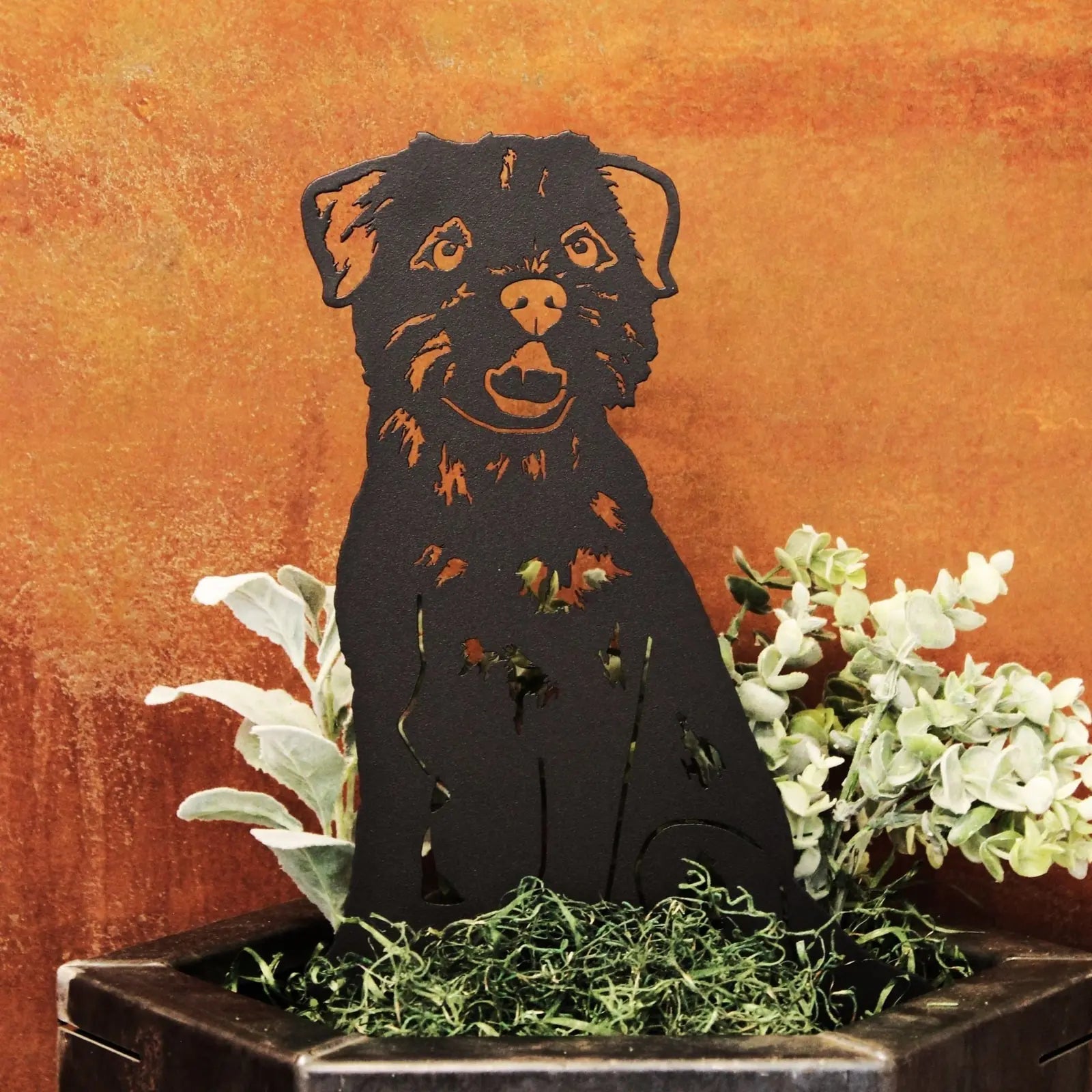 Black metal silhouette of a Border Terrier sitting among green foliage in a decorative pot against a warm background.