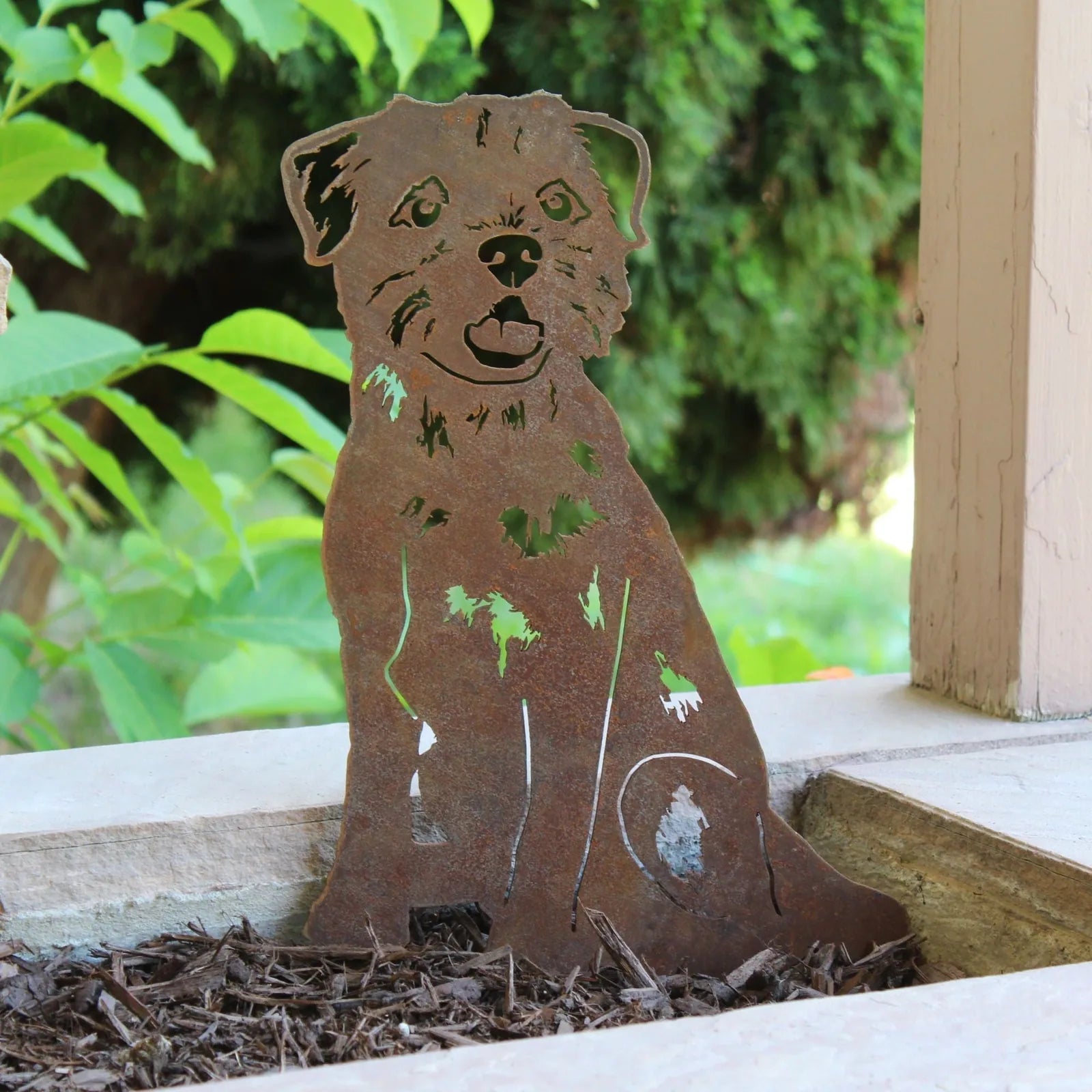 Rusty metal silhouette of a Border Terrier sitting, set against green foliage, perfect for outdoor decor.