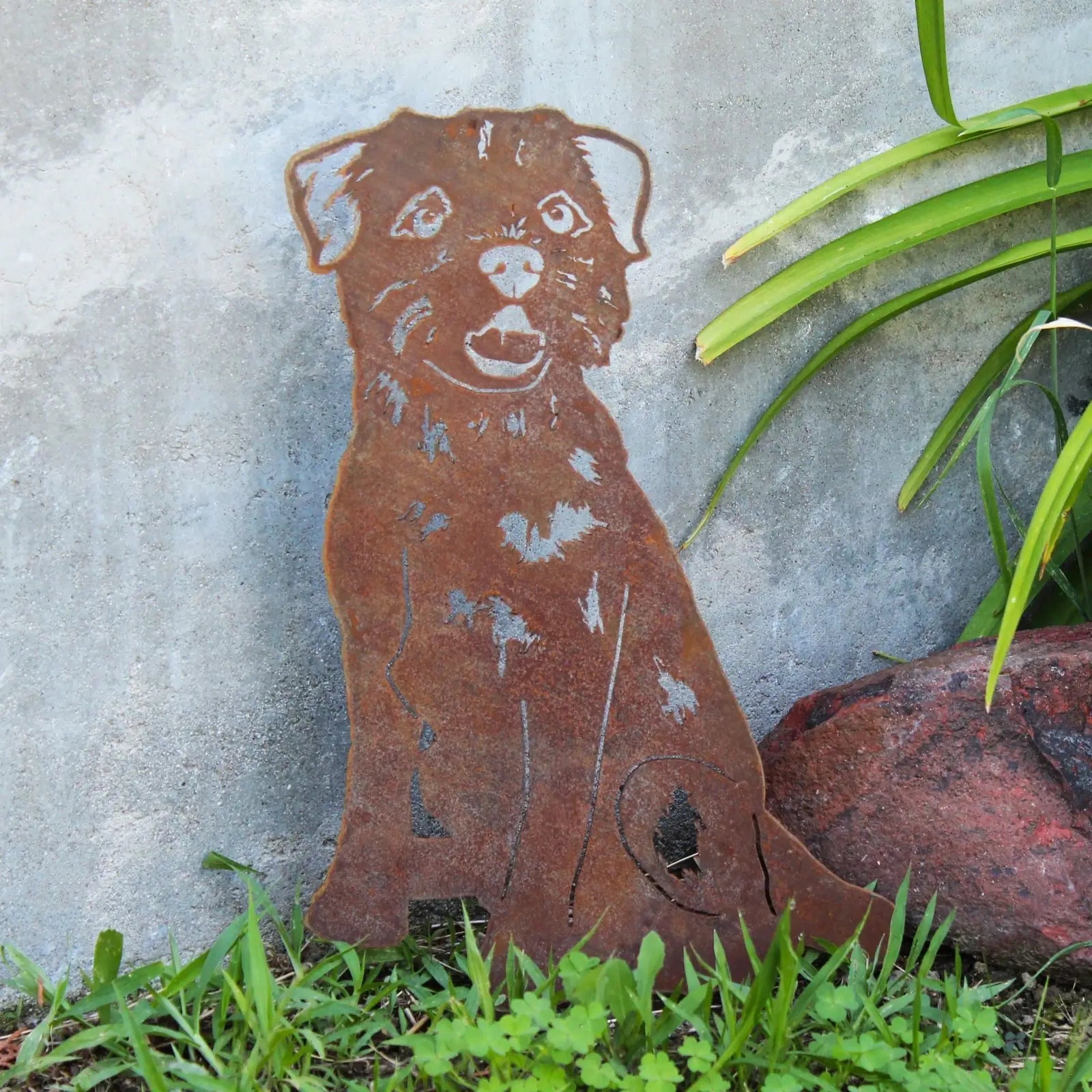 Handmade Corten steel Border Terrier silhouette, rusted finish, set in grass near a decorative stone and greenery.
