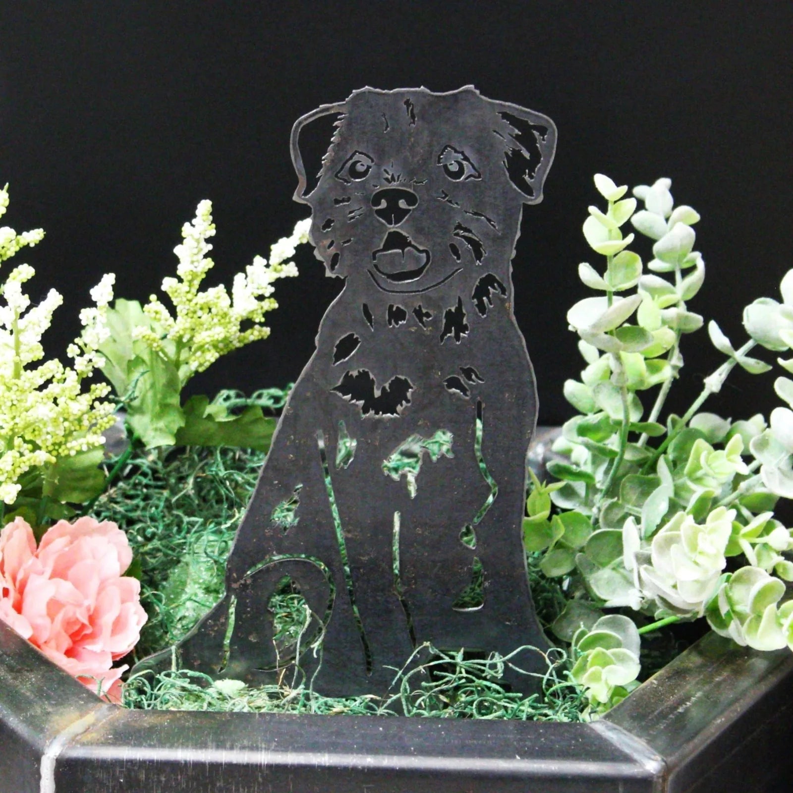 Black Border Terrier metal silhouette displayed among green foliage and decorative flowers in a planter.