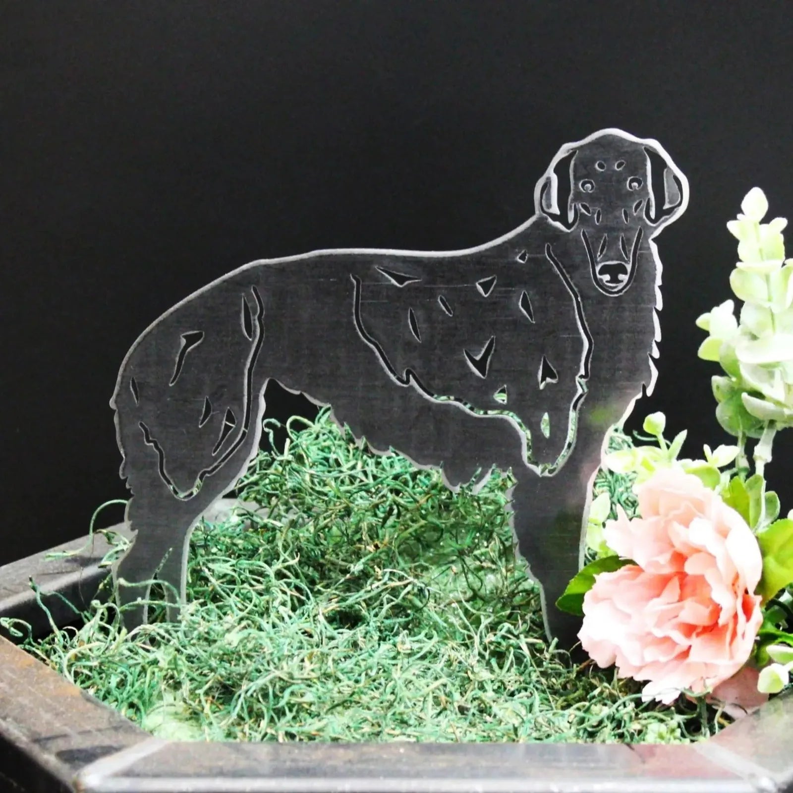 Borzoi dog silhouette in sleek metal, resting on green moss with a pink flower and foliage, perfect for outdoor decor.