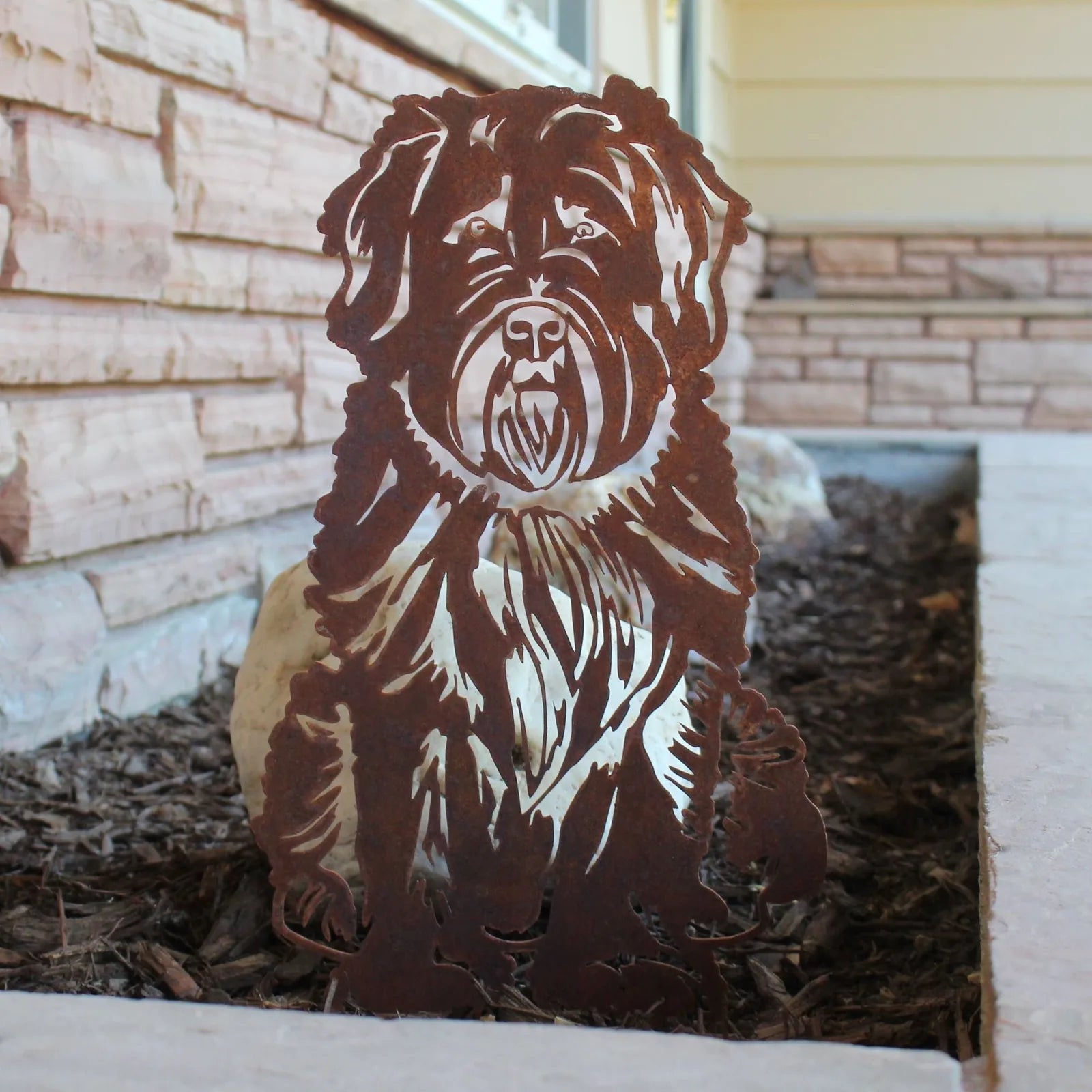 Bouvier Des Flandres metal silhouette in weathered Corten steel, positioned in garden mulch with stone backdrop.