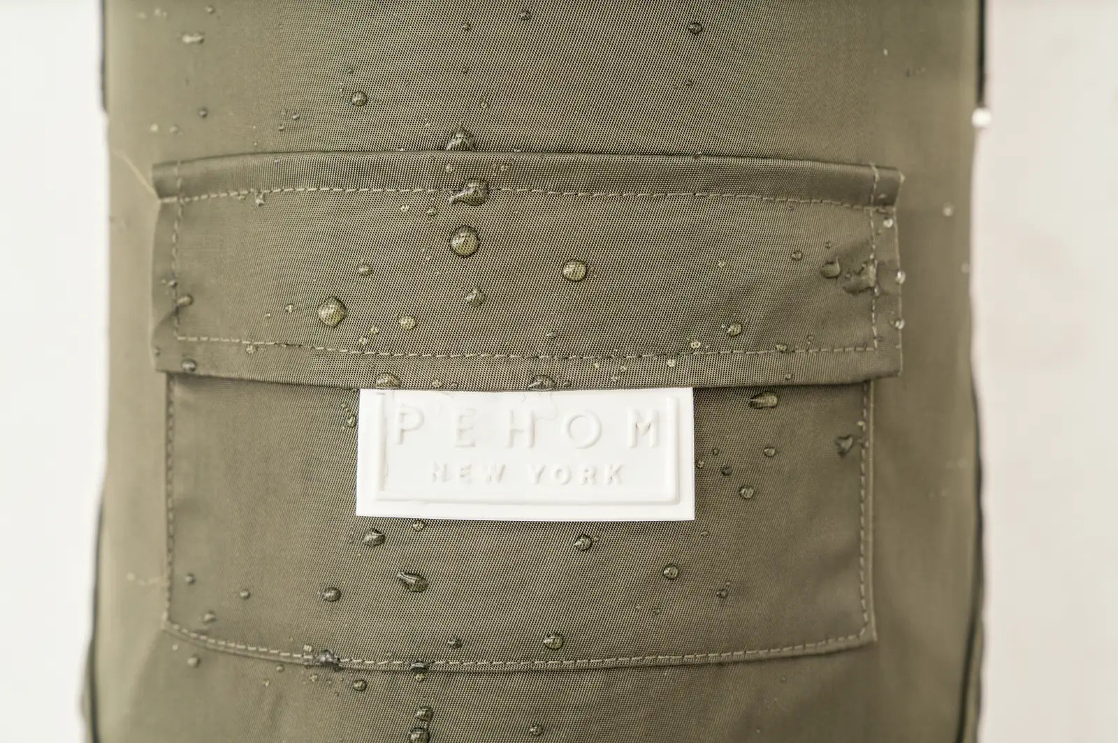 Beige Bowery Raincoat by pehomnyc with water droplets and logo patch, showcasing its water-resistant design.