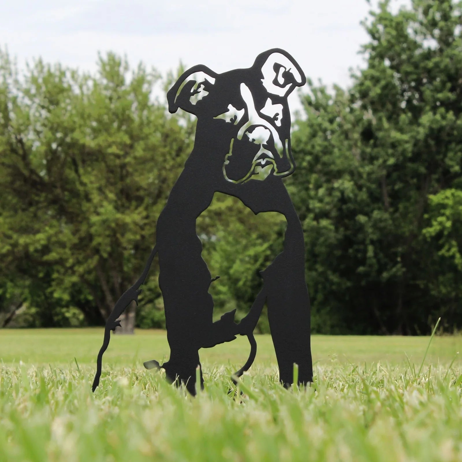 Boxer silhouette sculpted from black metal stands in grass, surrounded by greenery, ideal for outdoor dog decor.