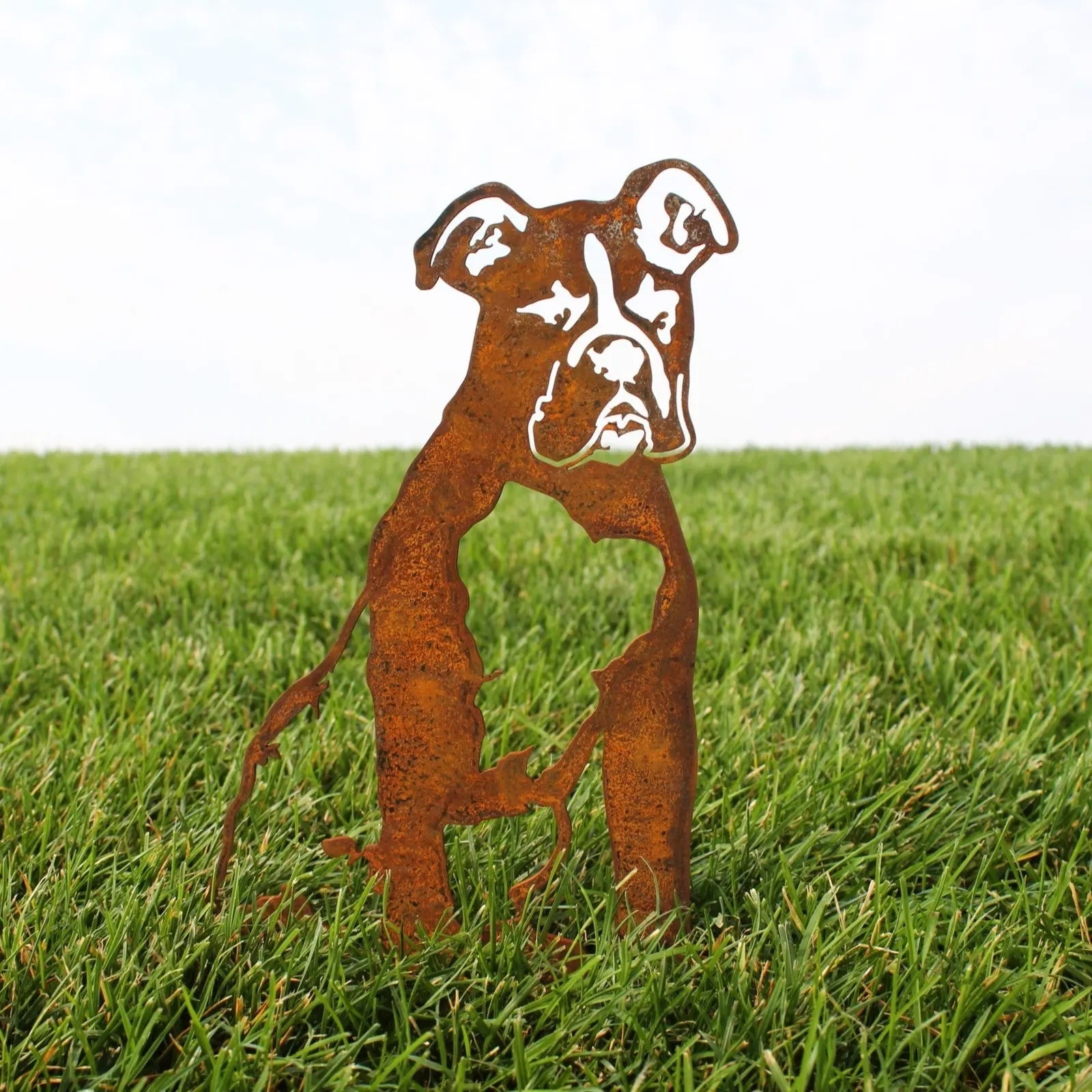 Boxer metal silhouette in rusted Corten steel, standing on green grass with a cloudy sky backdrop.