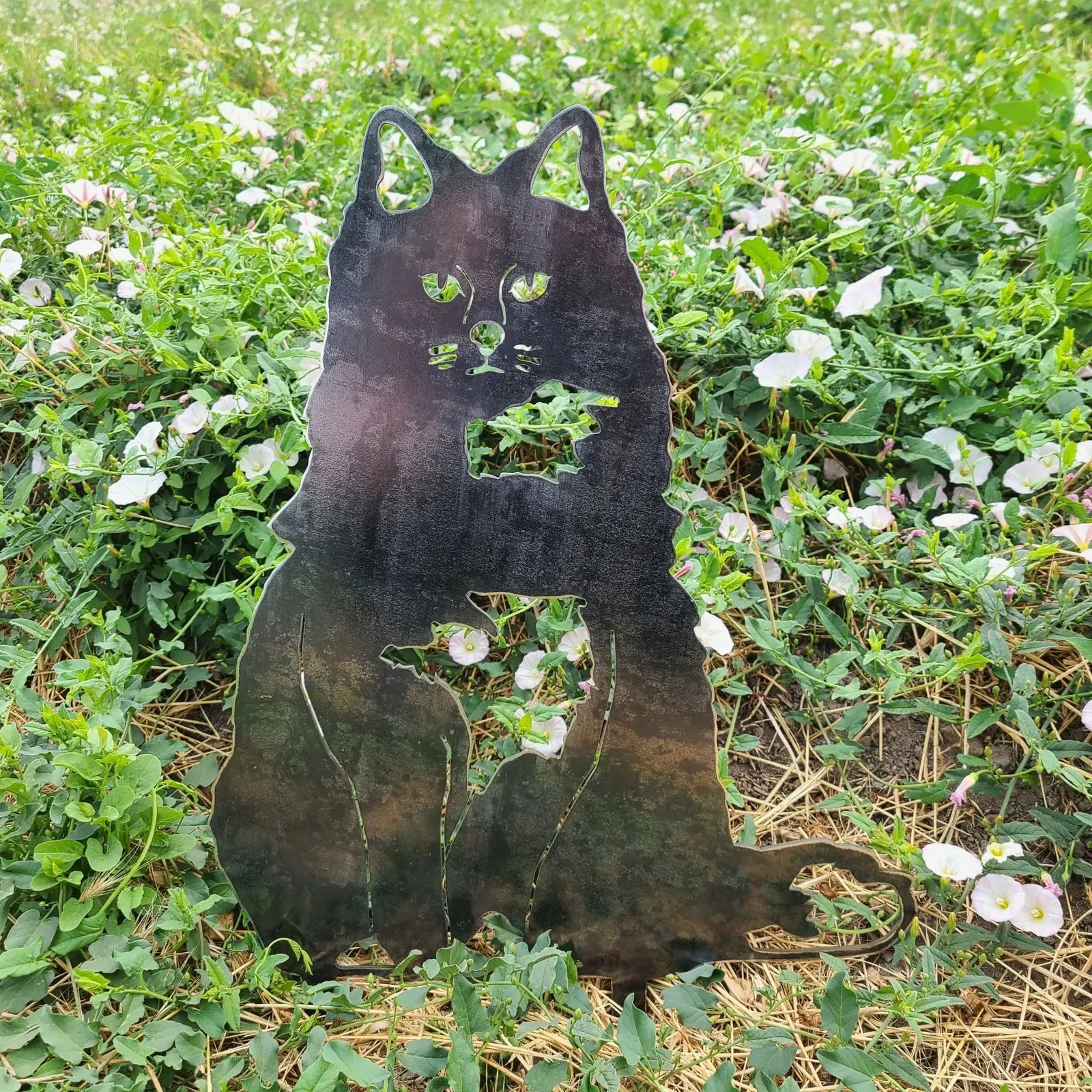 Handmade black metal silhouette of a British Longhair cat among green grass and pink flowers.