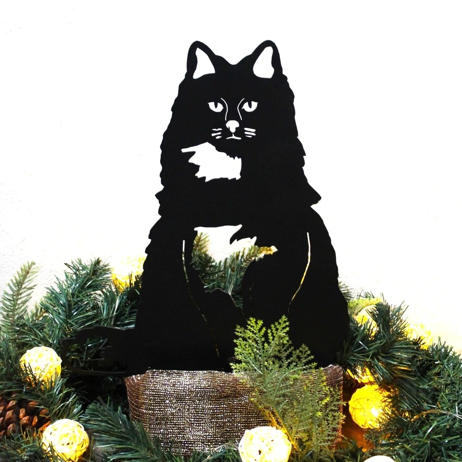 Black metal silhouette of a British Longhair cat, set among festive greenery and decorative lights.