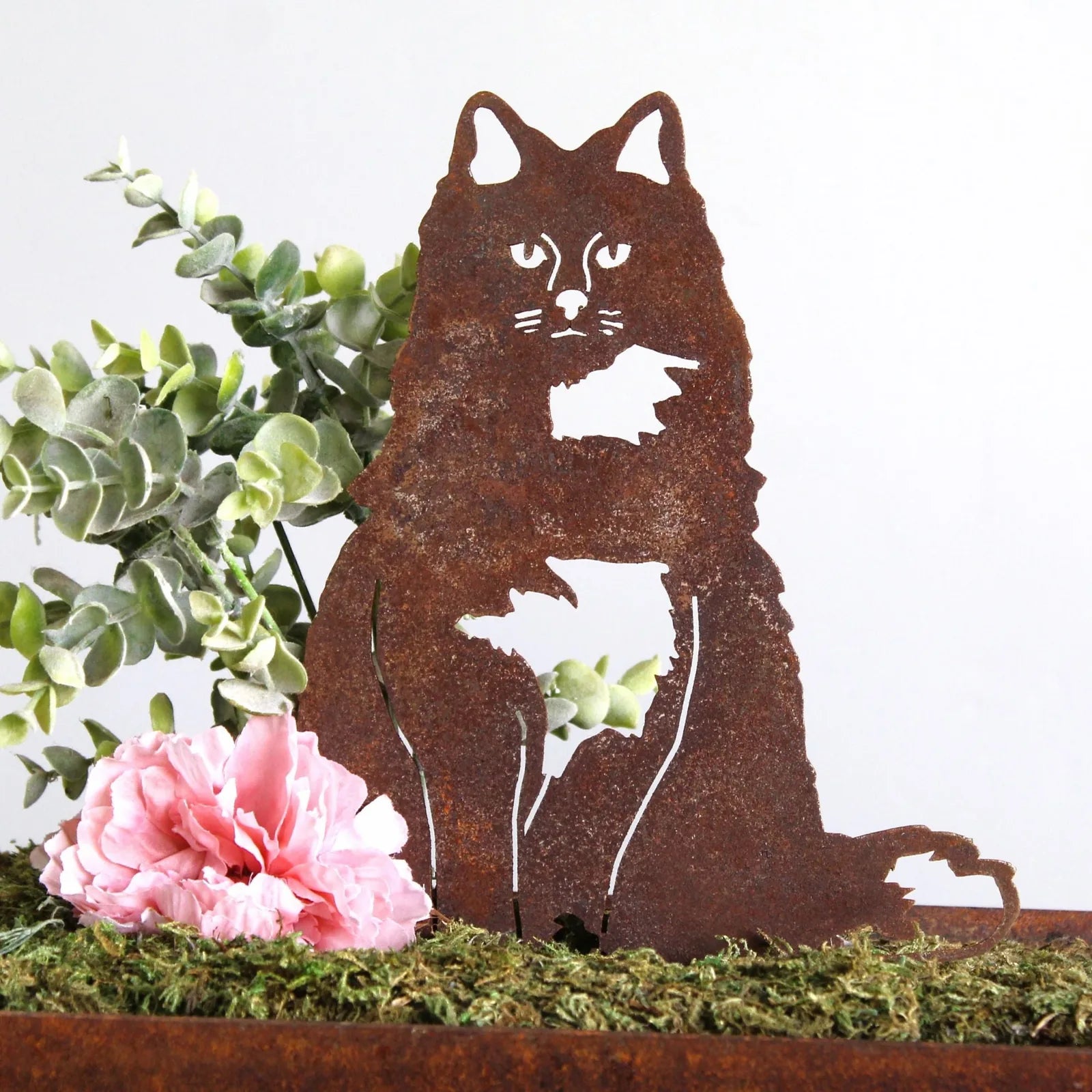 Handmade rust-colored metal silhouette of a British Longhair cat, surrounded by greenery and pink flowers.