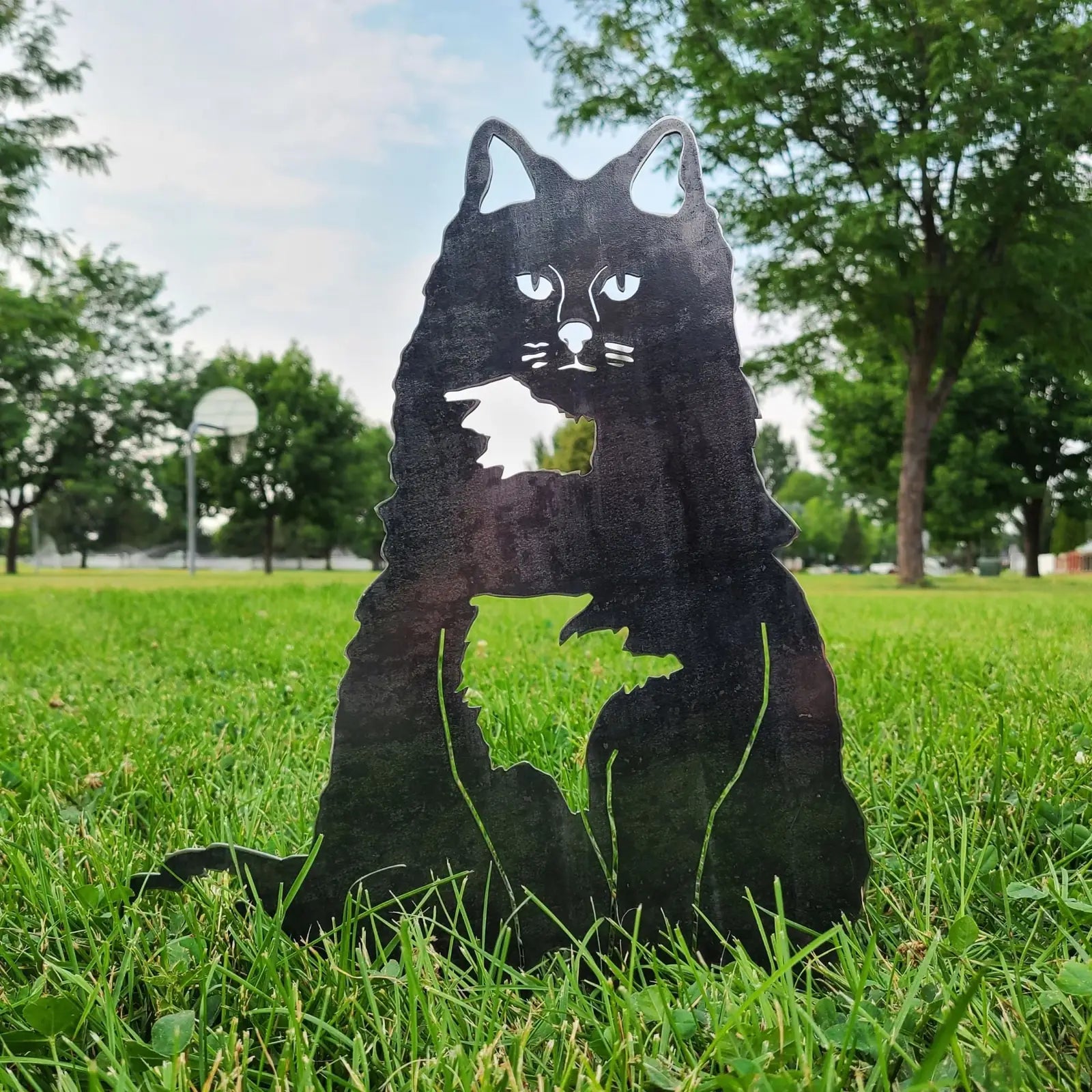 Handmade black metal silhouette of a British Longhair cat, set on grass with trees and a basketball court in the background.
