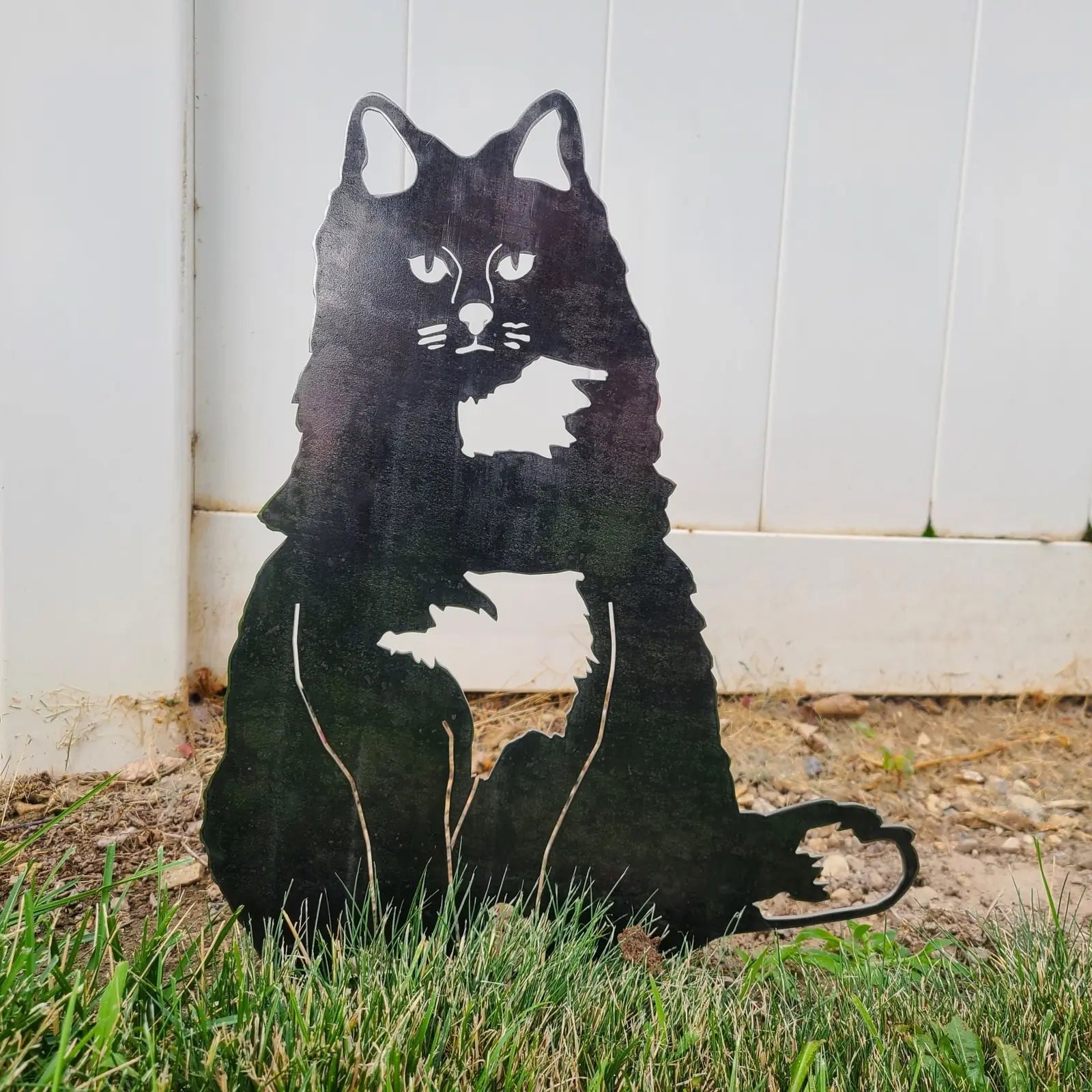 Handcrafted black metal silhouette of a British Longhair cat, displayed in grass against a white fence background.