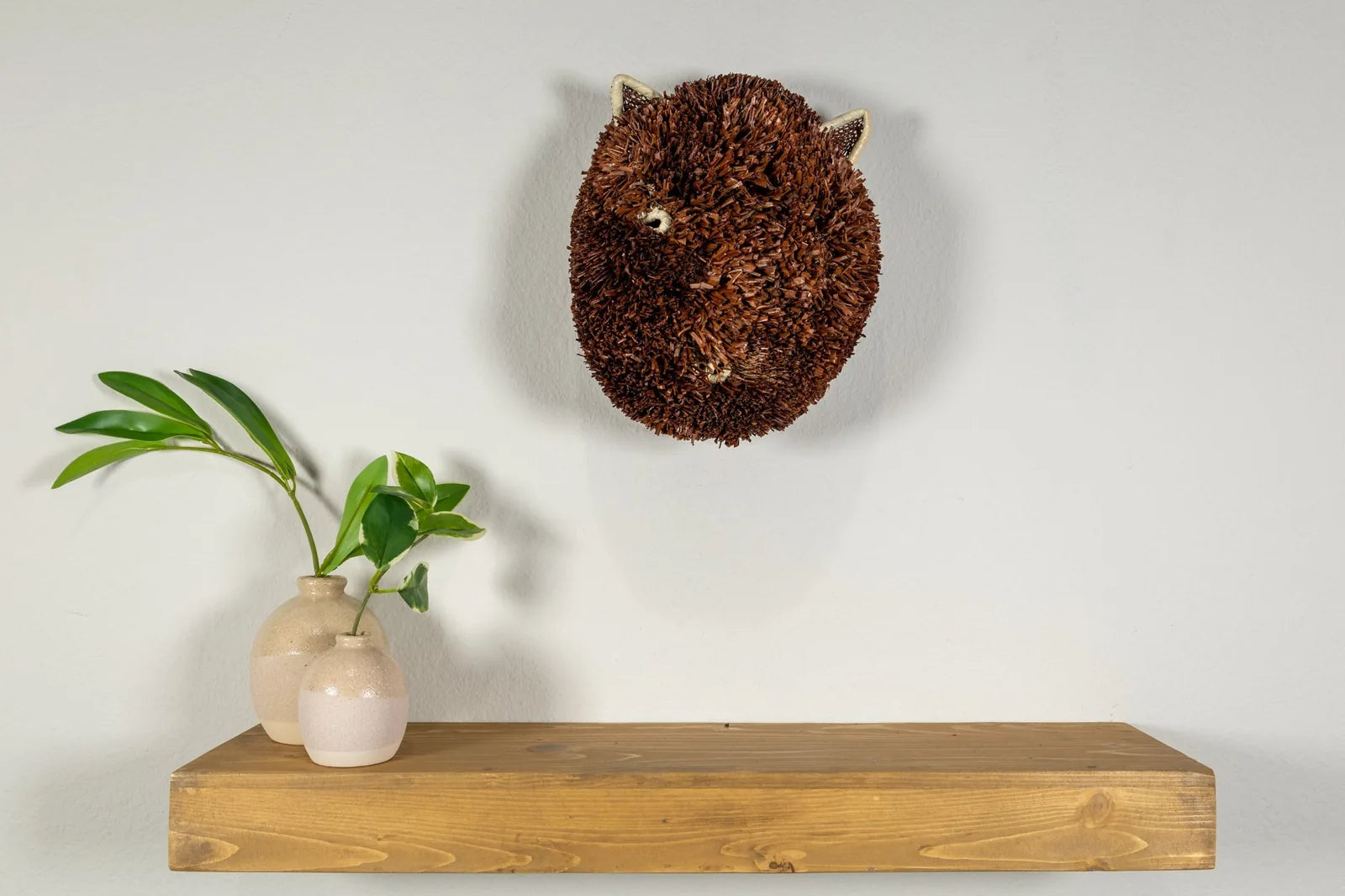 Handwoven brown and white Labradoodle dog mask, crafted from natural materials, displayed on a wooden shelf with plants.