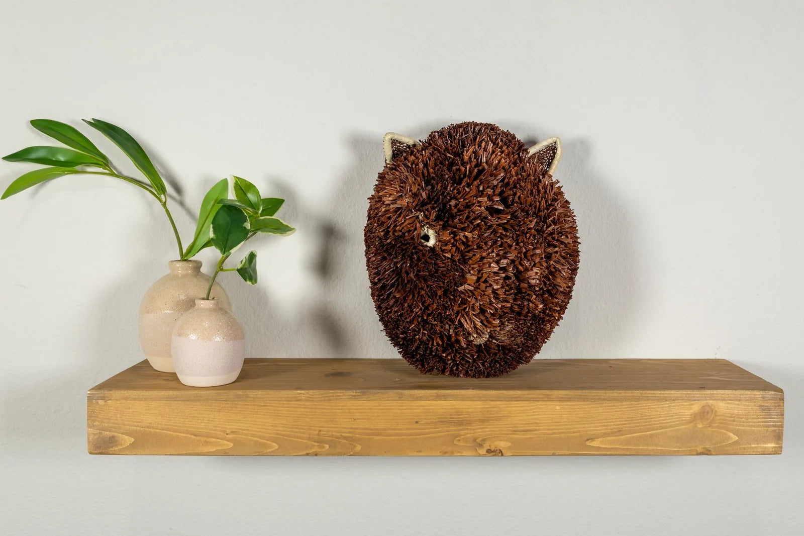 Handwoven brown and white Labradoodle dog mask displayed on a wooden shelf with two vases and greenery.
