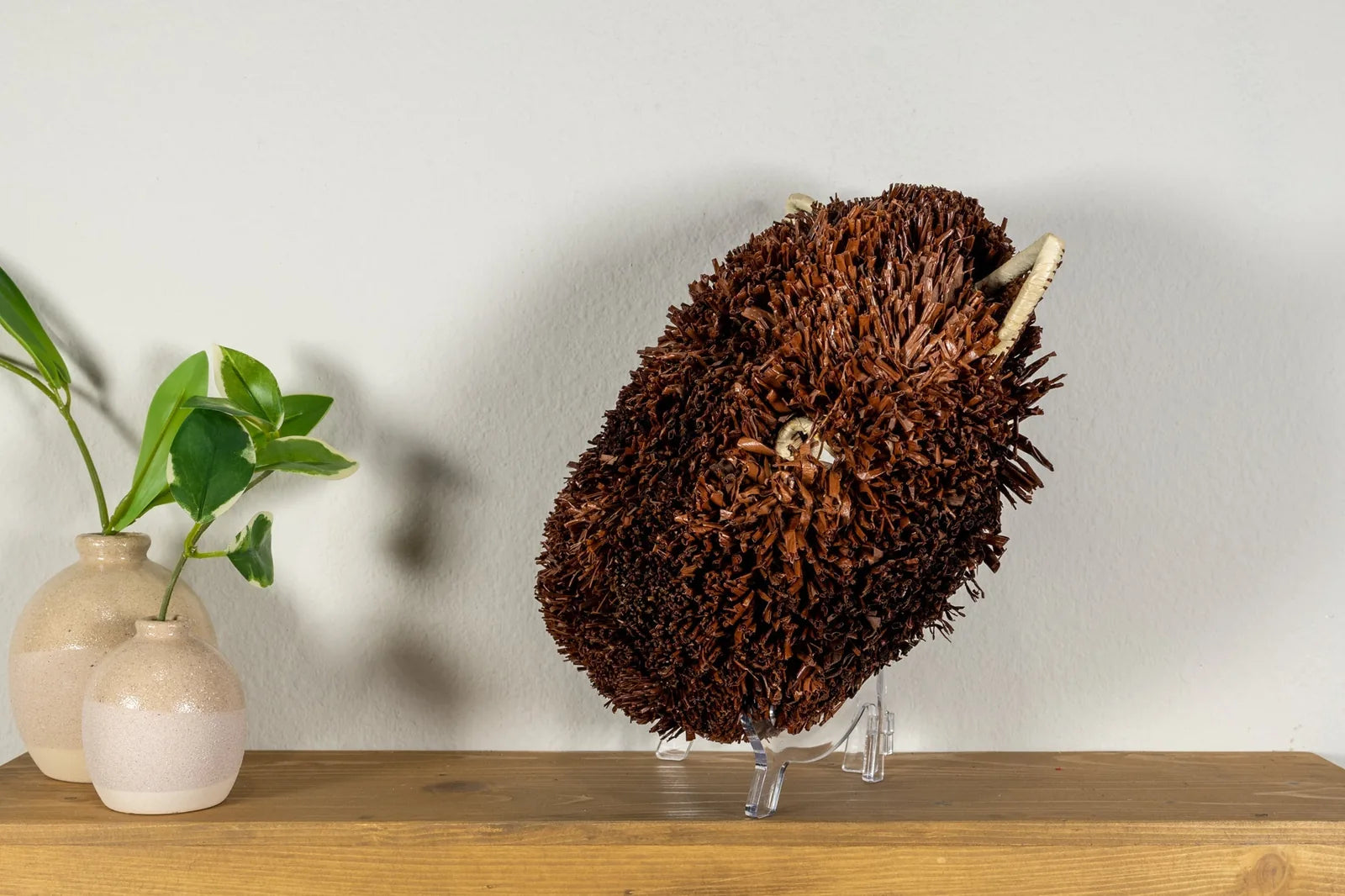 Handwoven brown and white Labradoodle dog mask displayed on a wooden surface with decorative vases and greenery.