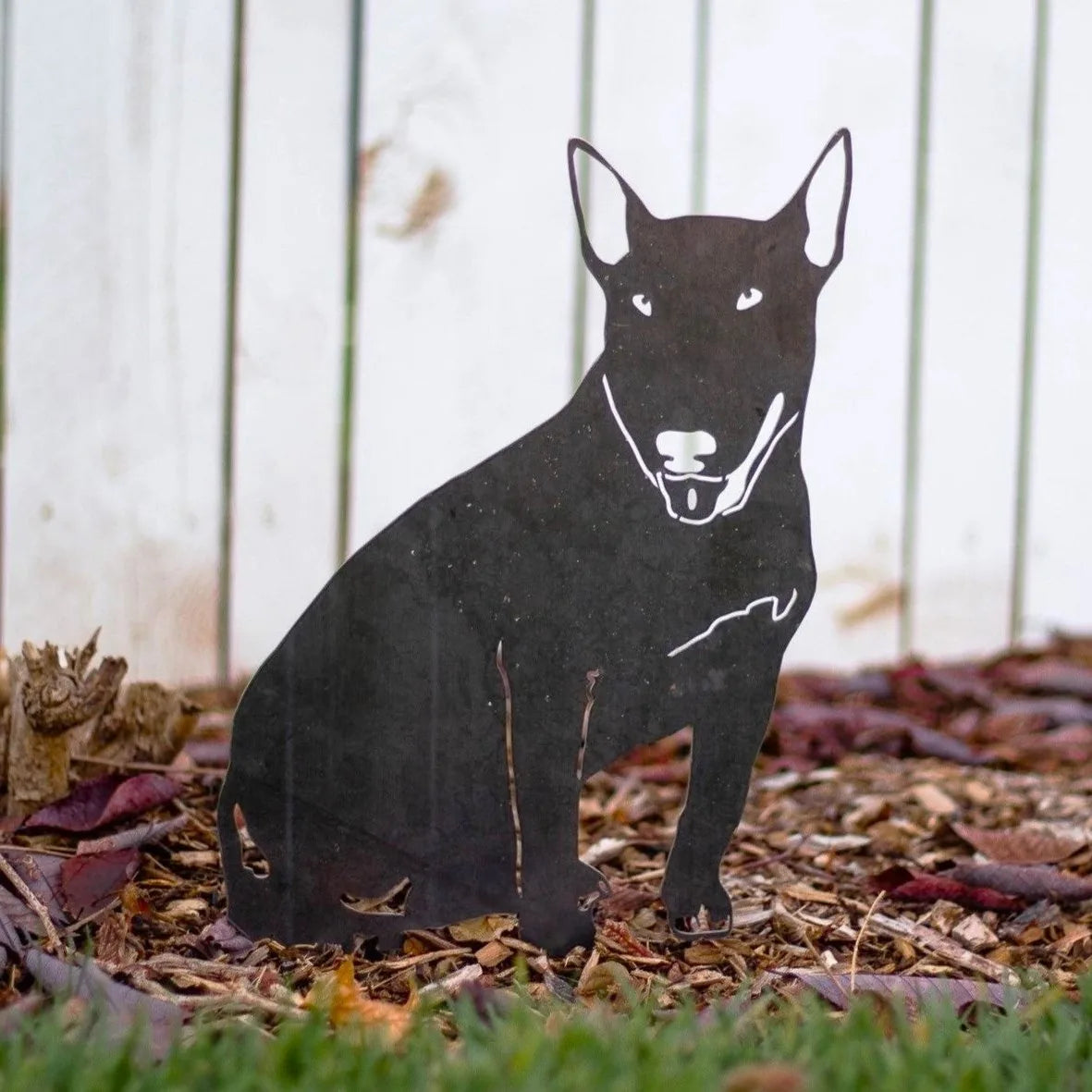 Black Bull Terrier metal silhouette resting among mulch, showcasing its detailed design against a wooden fence.