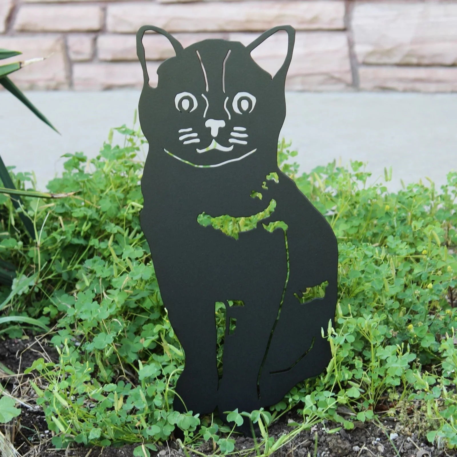 Black metal silhouette of a cat sits among green plants, showcasing intricate cut-out details and a smooth, matte finish.