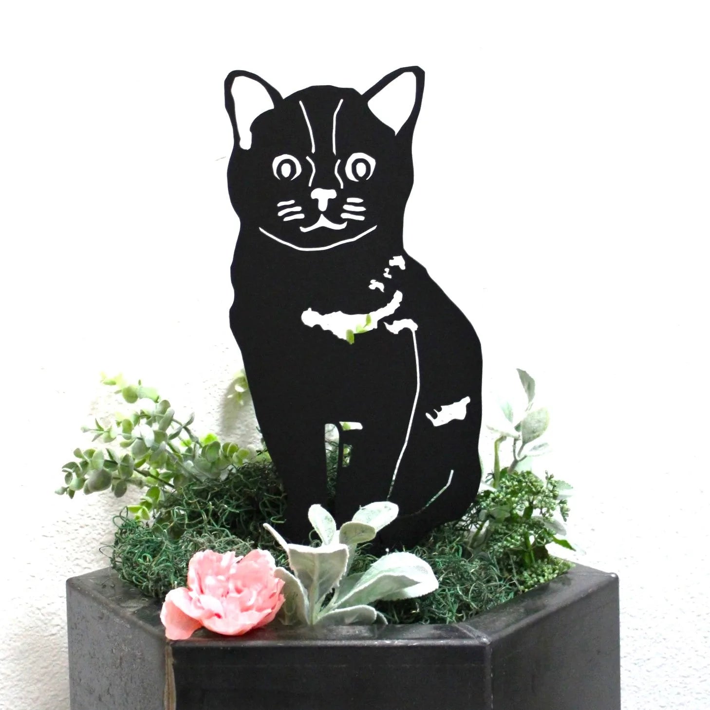 Black metal silhouette of a cat, nestled among greenery and a pink flower in a decorative planter.