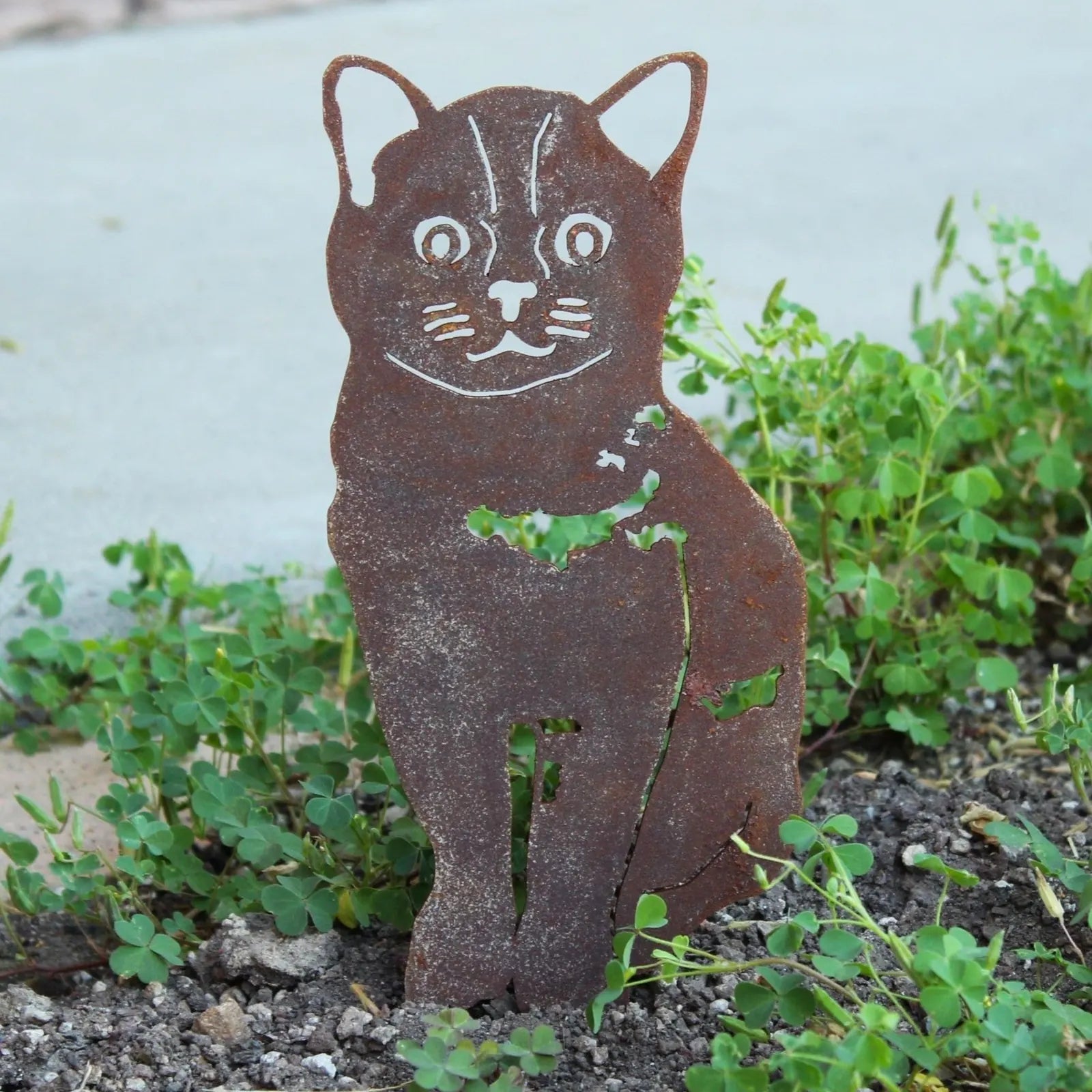 Handmade Burmese metal silhouette in weathered Corten steel, depicting a cat, surrounded by green ground cover.