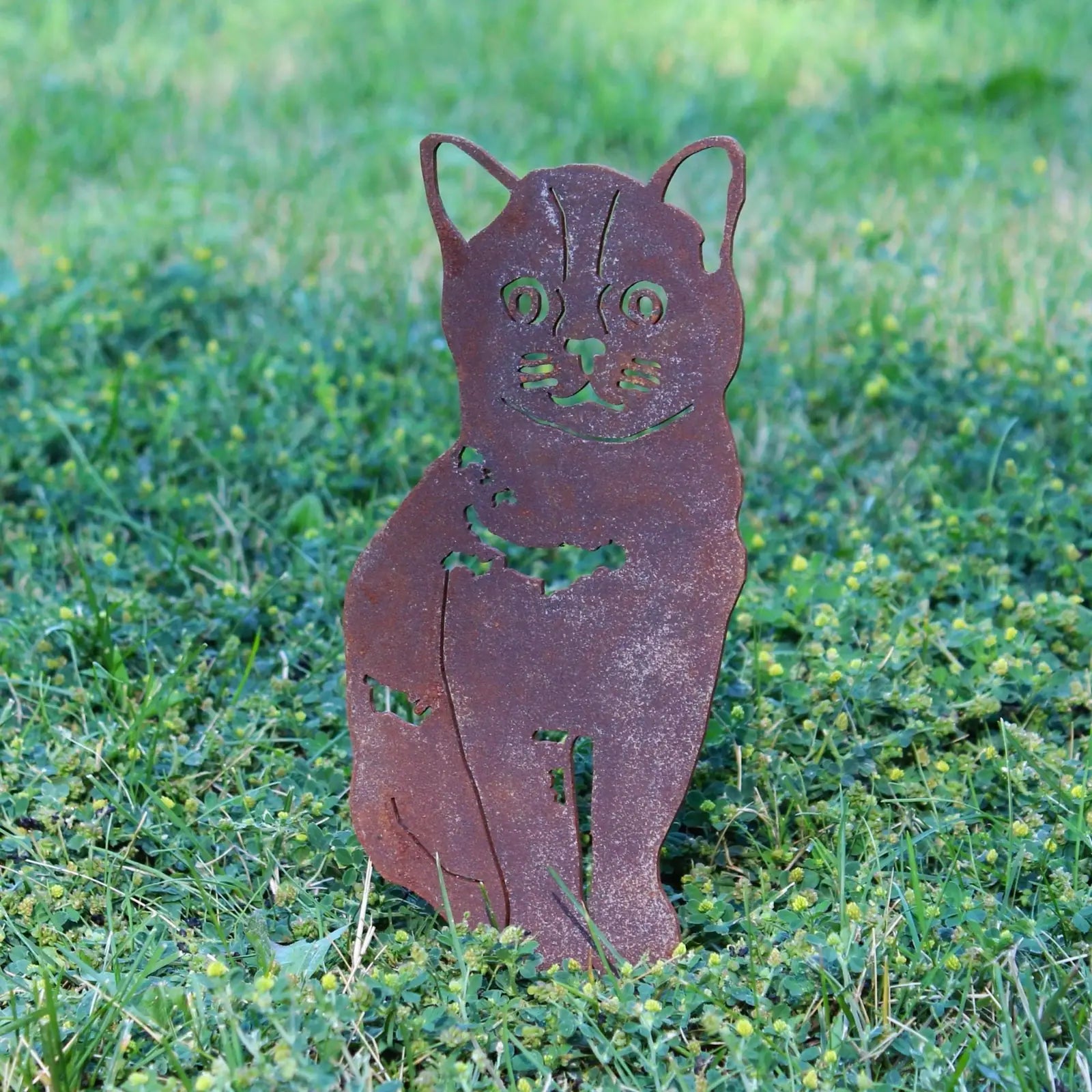 Rusty Corten steel silhouette of a sitting cat amidst green grass and small yellow clover flowers.
