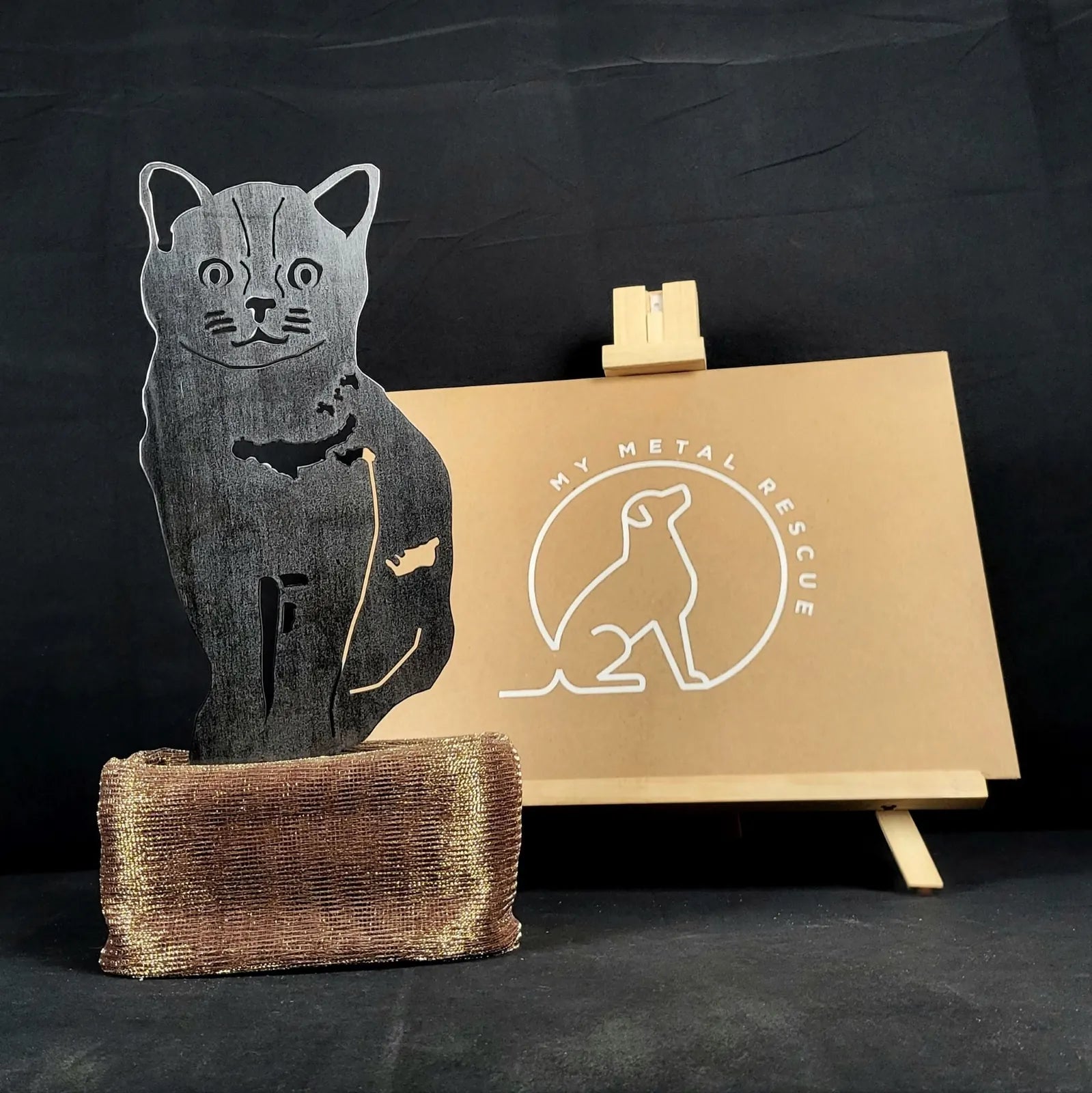 Burmese cat metal silhouette in matte black, displayed on a textured brown base, with a branded sign in the background.