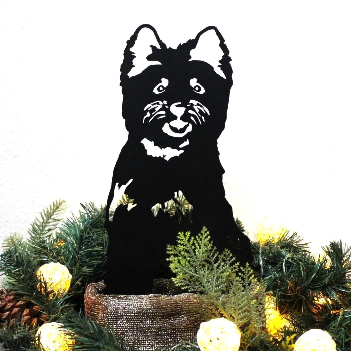Black metal silhouette of a Cairn Terrier, surrounded by greenery and soft lights, showcasing handmade craftsmanship.