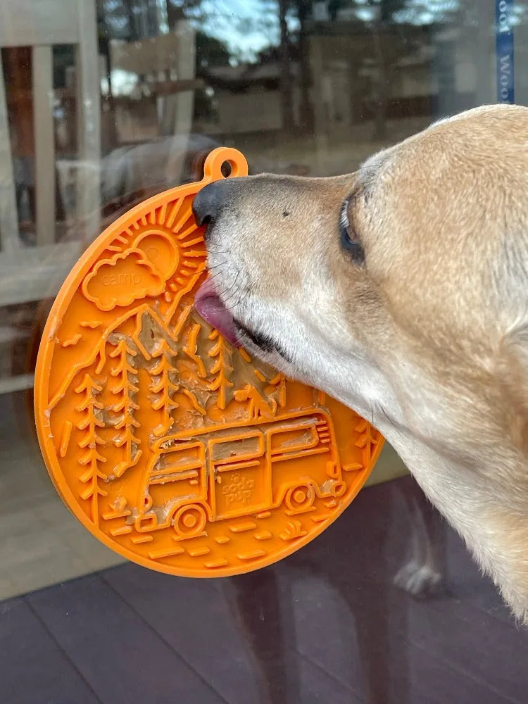 Dog licking a vibrant orange Camp eMat with retro camper van design and textured surface, secured to a glass window.