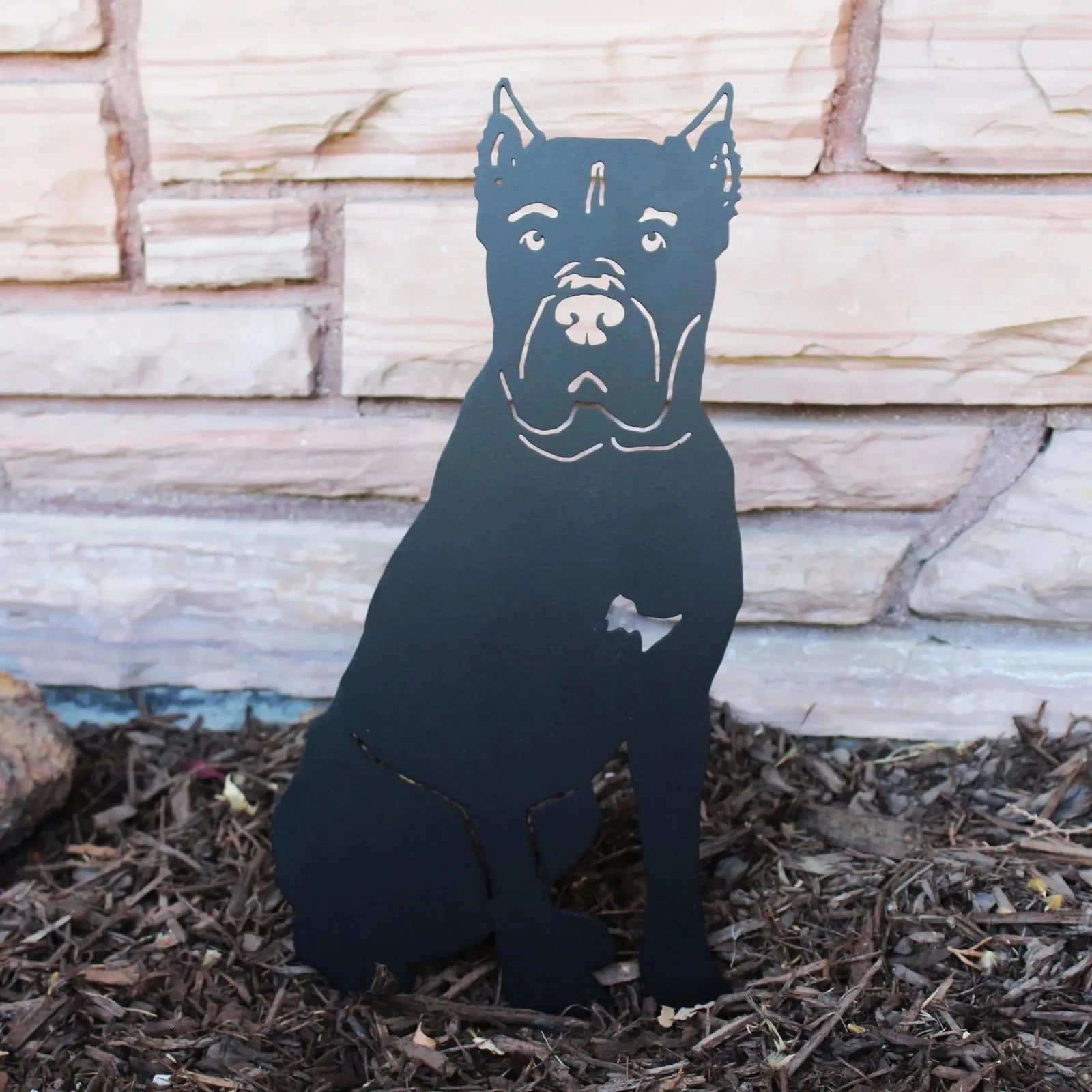 Cane Corso metal silhouette in matte black, seated on mulch against a stone wall, adds durable dog decor to outdoor spaces.