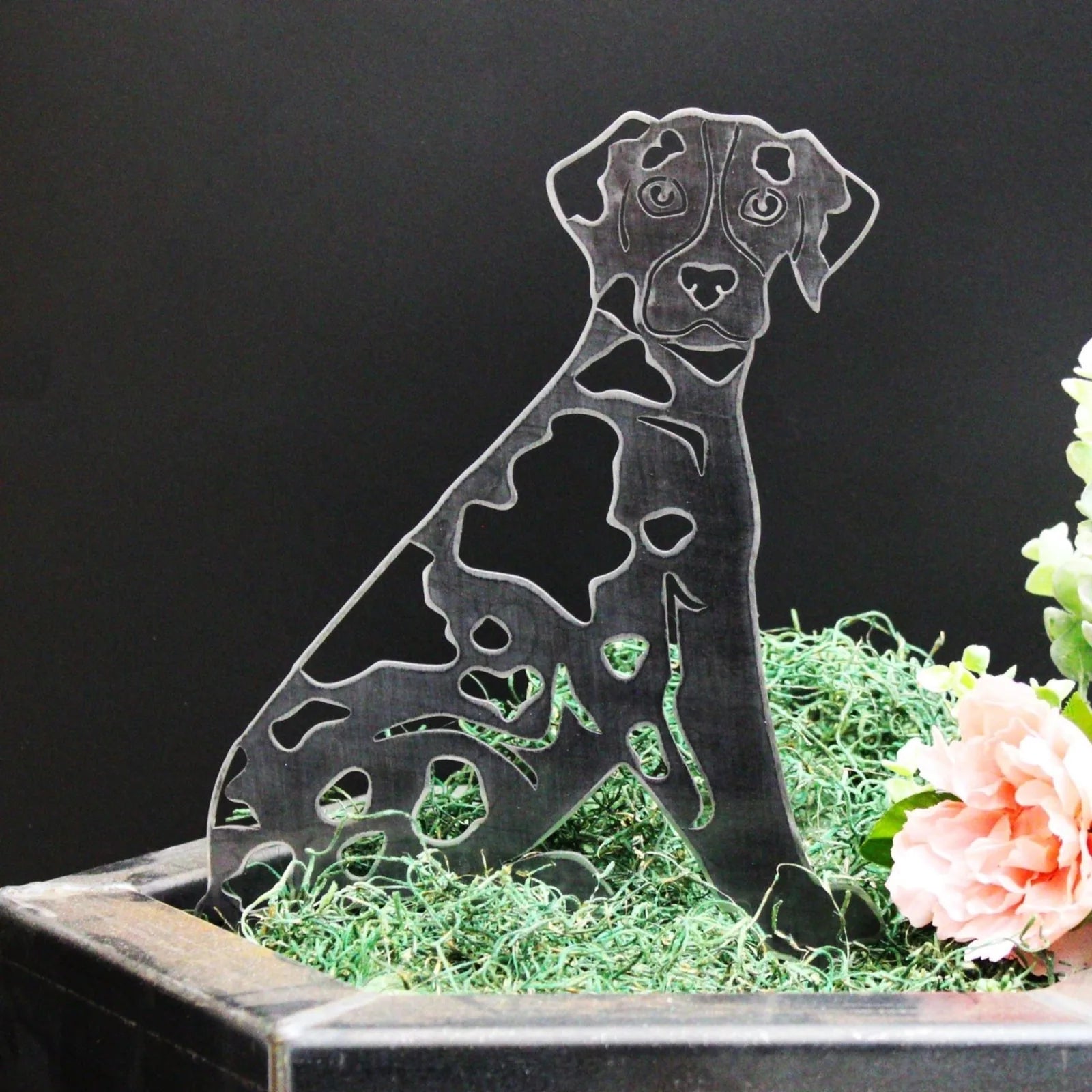Catahoula Leopard Dog silhouette in matte black metal, showcasing cutout patterns, set among green moss and flowers.