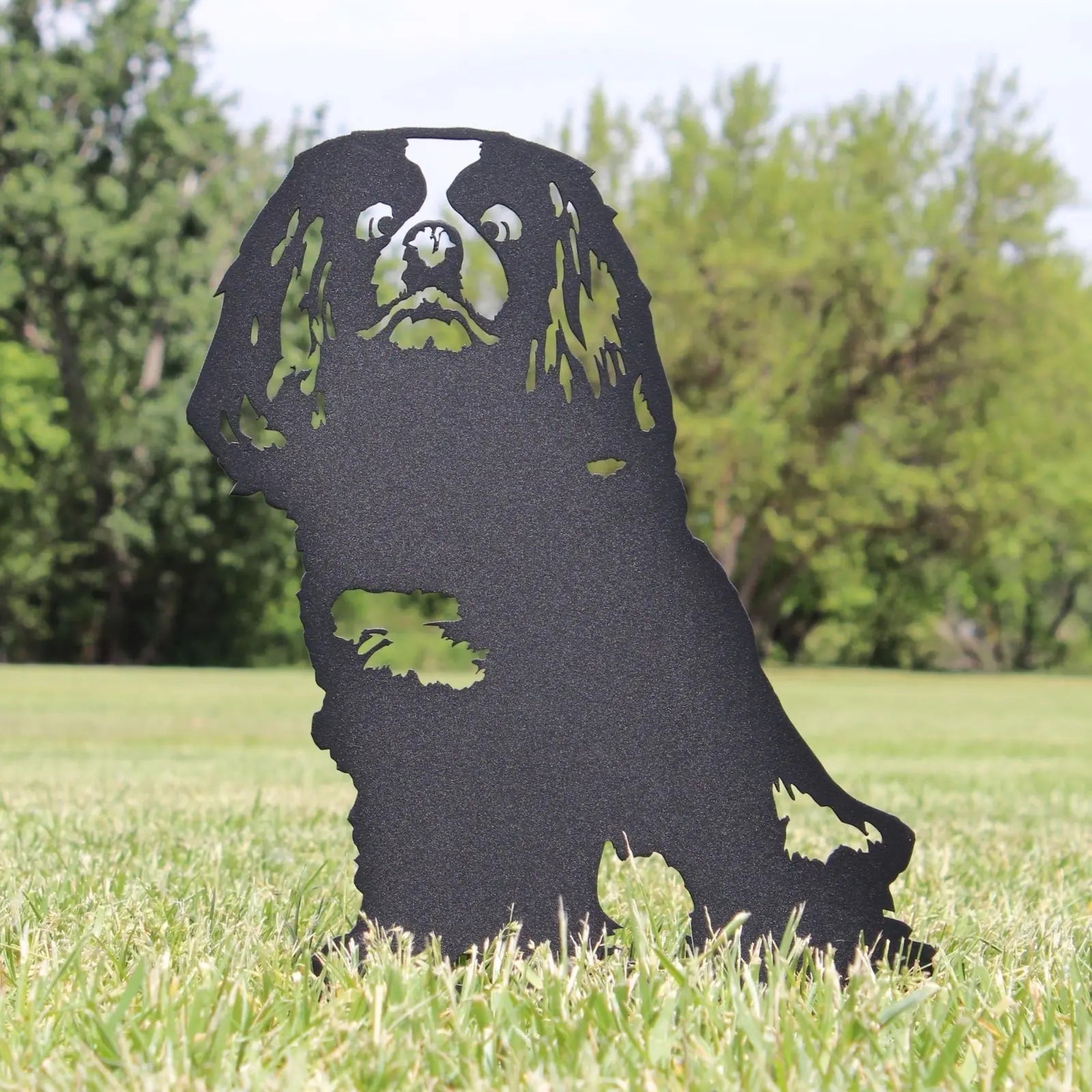 Cavalier King Charles Spaniel silhouette in black metal, set against a grassy outdoor backdrop.