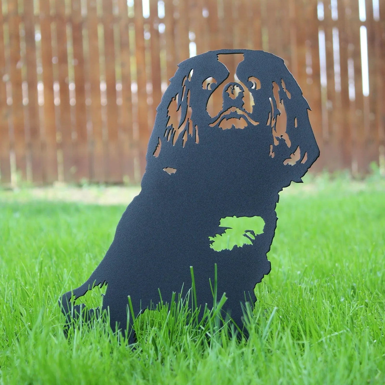 Cavalier King Charles Spaniel silhouette in matte black metal, standing on green grass against a wooden fence backdrop.