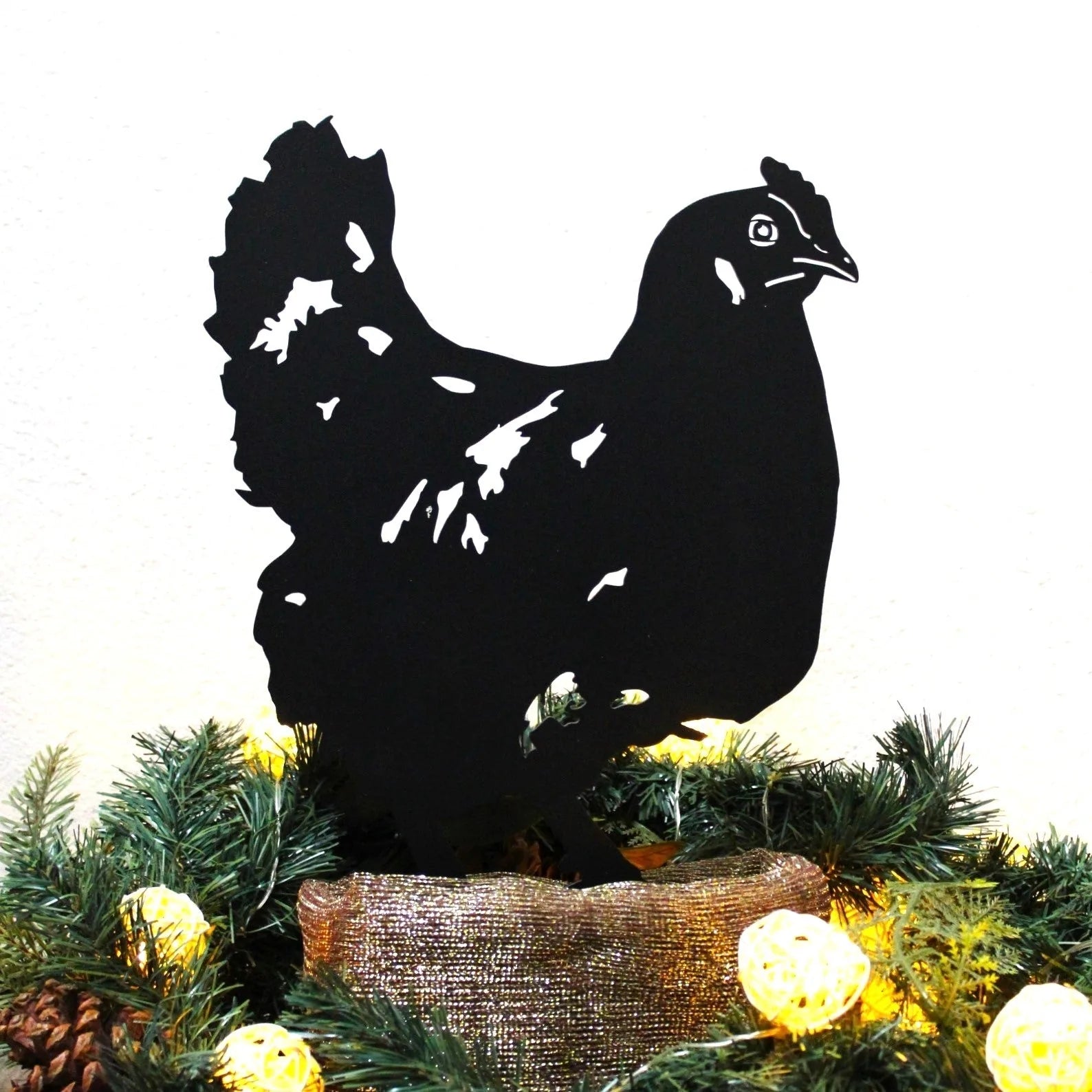 Black chicken silhouette made from metal, showcasing intricate details, positioned among greenery and decorative lights.