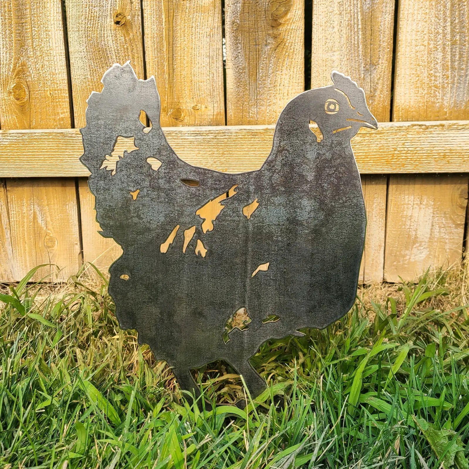 Rustic chicken silhouette in weathered Corten steel, set against a wooden fence and surrounded by green grass.