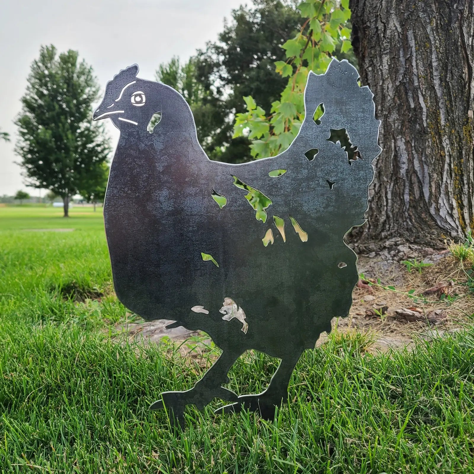 Corten steel chicken silhouette with cut-out details, set in lush green grass and surrounded by trees.