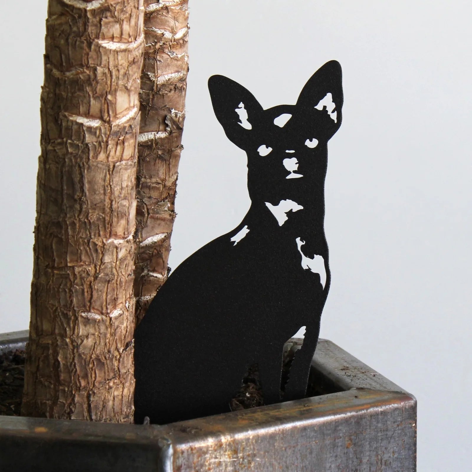 Black metal silhouette of a Chihuahua dog nestled beside a tree trunk in a planter. Ideal for outdoor decor.