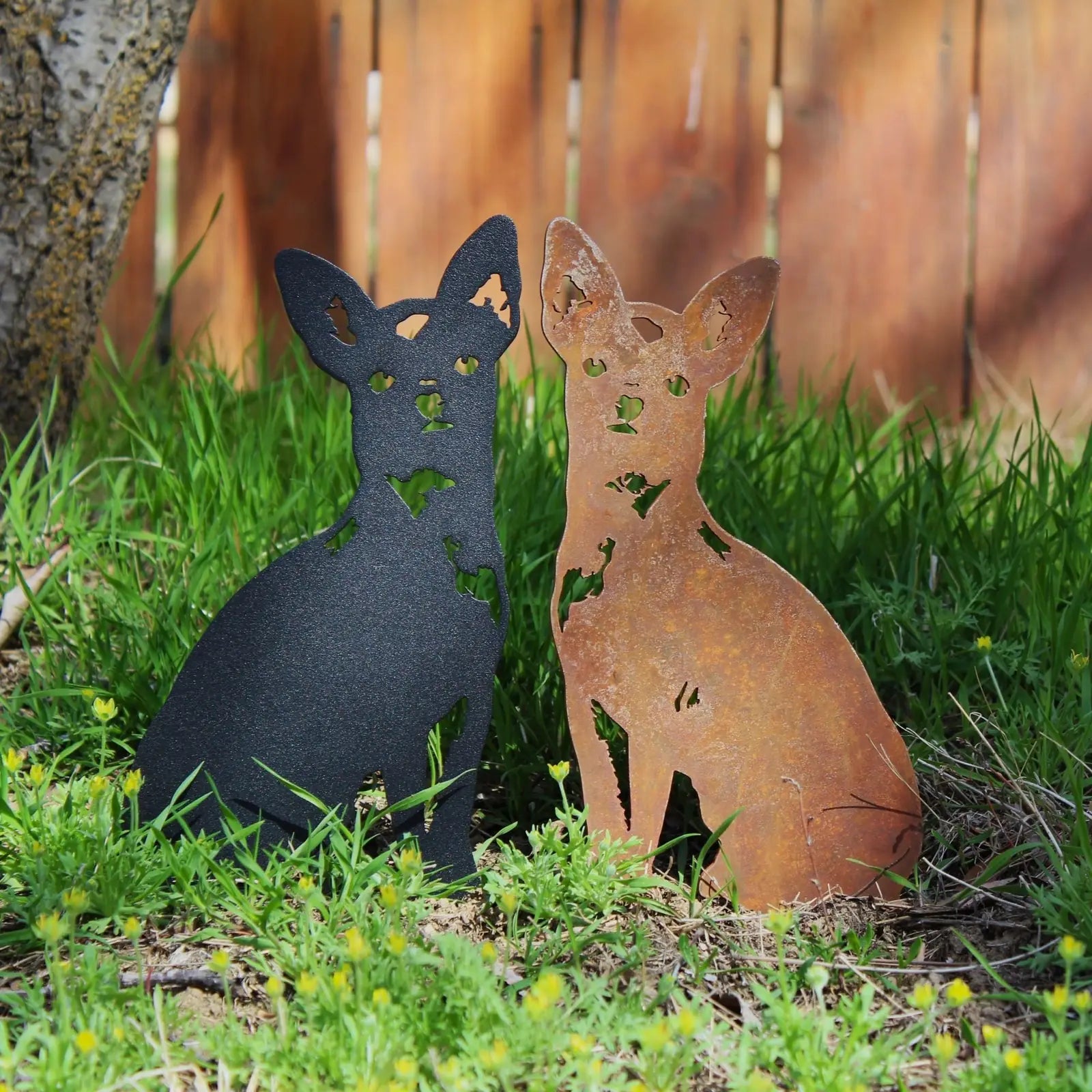 Black and rust metal silhouettes of seated Chihuahuas on green grass, surrounded by wildflowers and a wooden backdrop.
