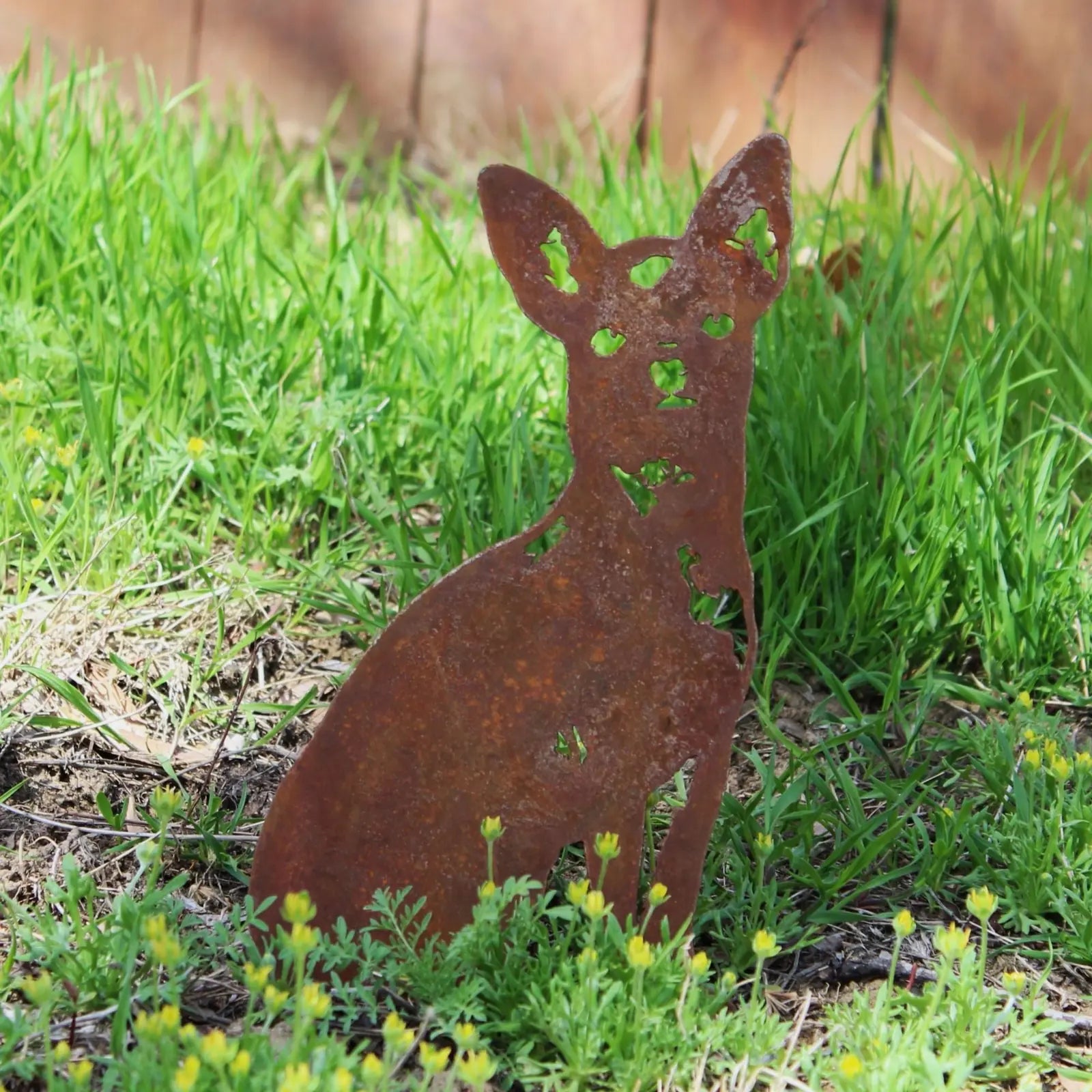 Rusty metal silhouette of a Chihuahua dog sitting among green grass and wildflowers, perfect for outdoor decor.