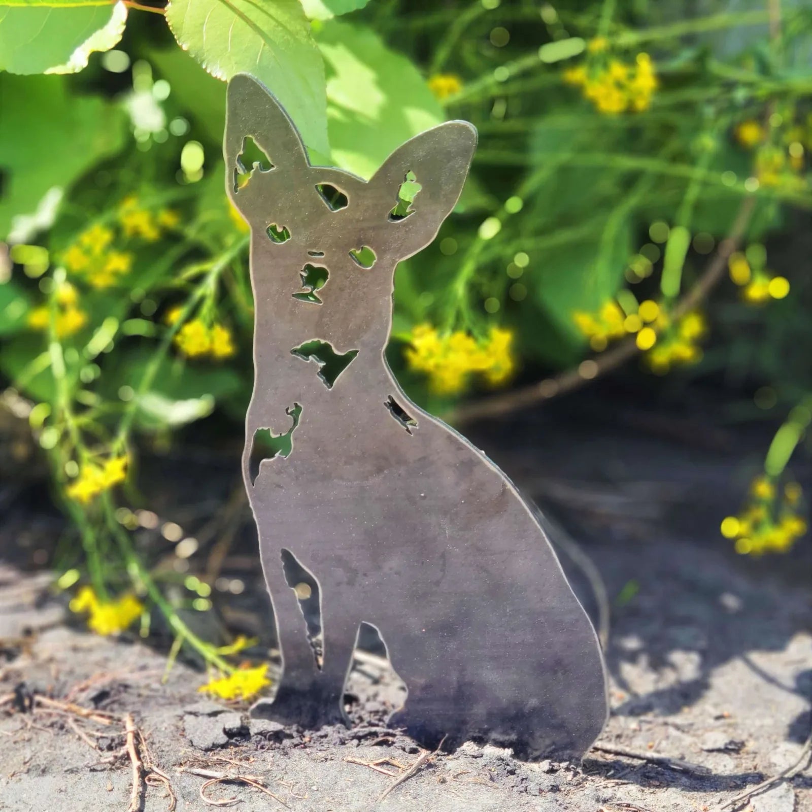 Handcrafted metal silhouette of a Chihuahua in Corten steel, set against greenery and yellow flowers.