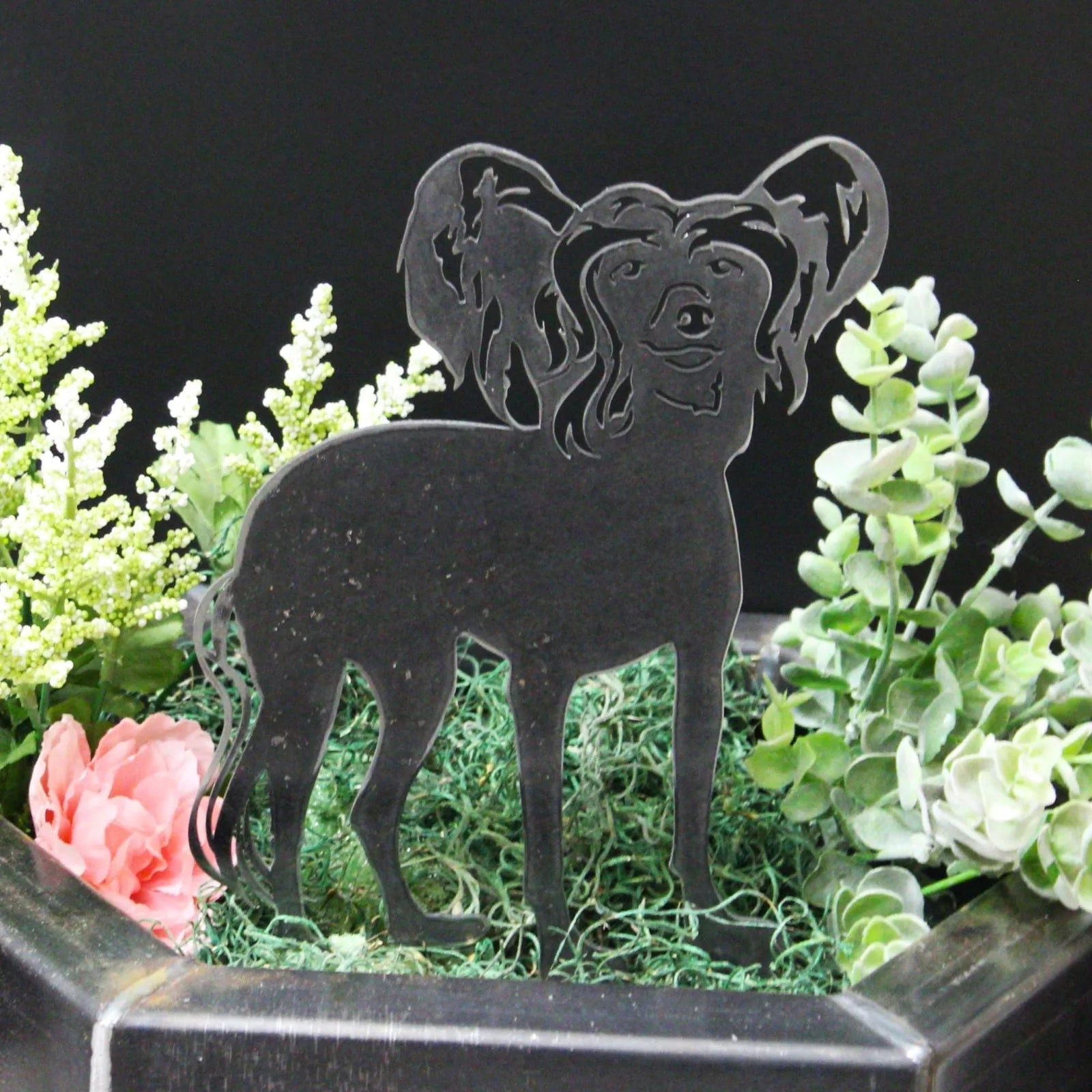 Black metal silhouette of a Chinese Crested dog among greenery in a decorative arrangement.