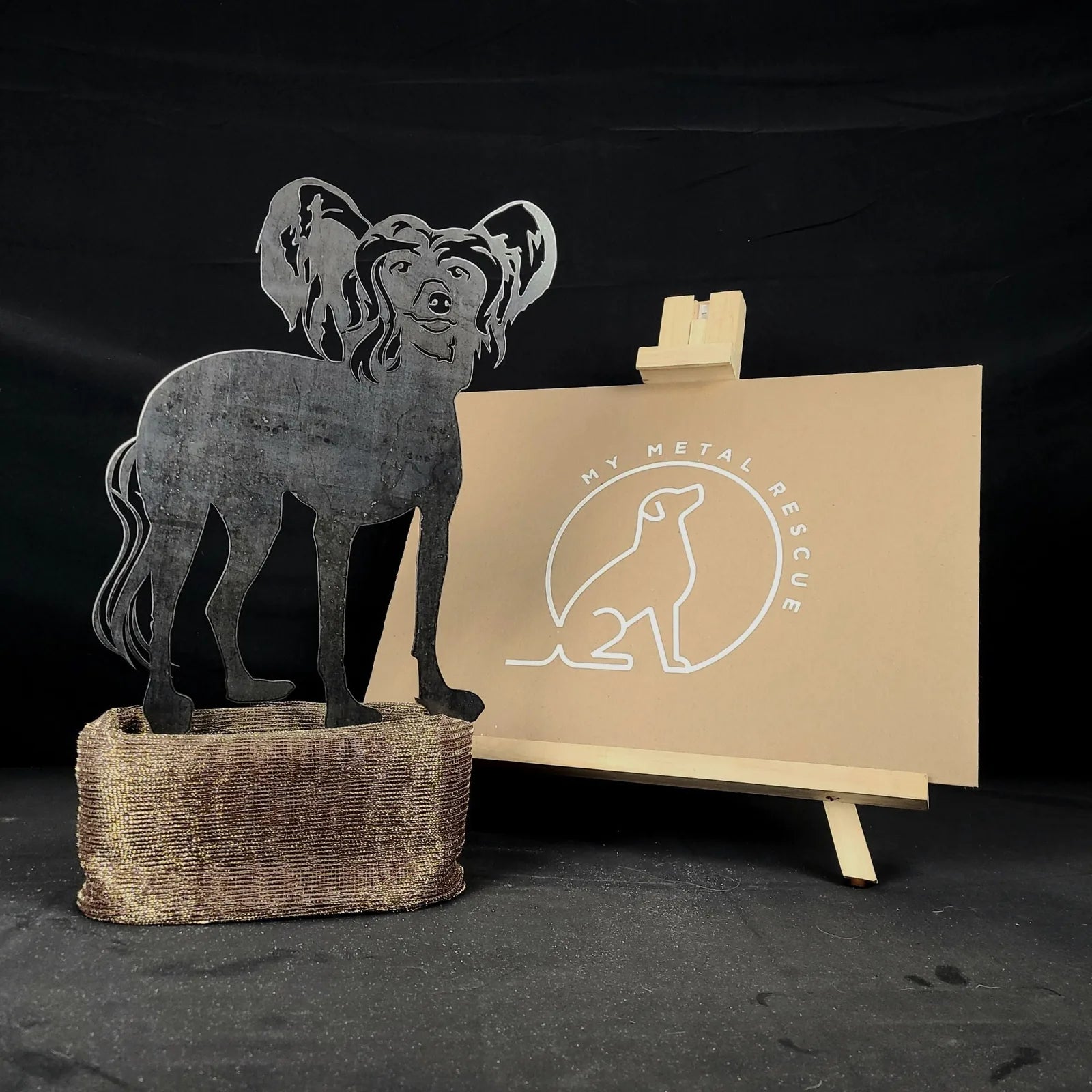 Handmade black metal silhouette of a Chinese Crested dog, displayed on a textured base with a logo backdrop.