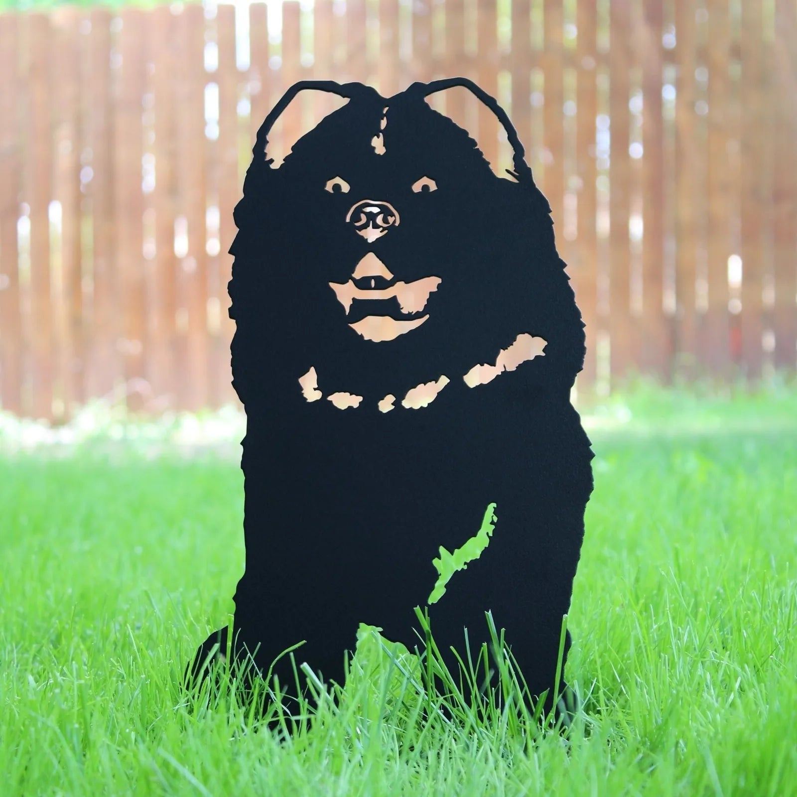 Chow Chow metal silhouette in matte black, standing on lush green grass with a wooden fence in the background.