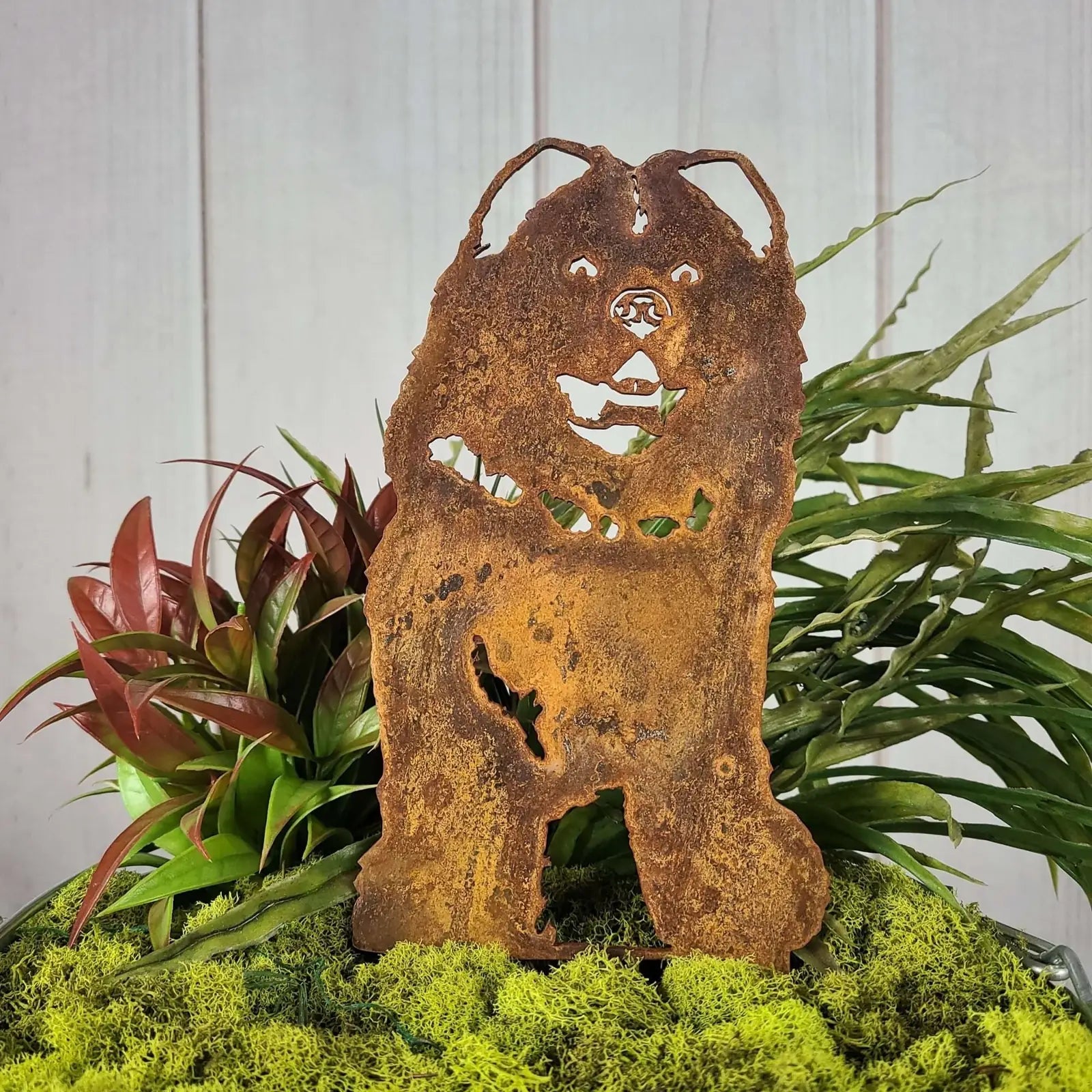 Rusty metal silhouette of a Chow Chow dog, surrounded by green plants and moss, showcasing outdoor decor charm.