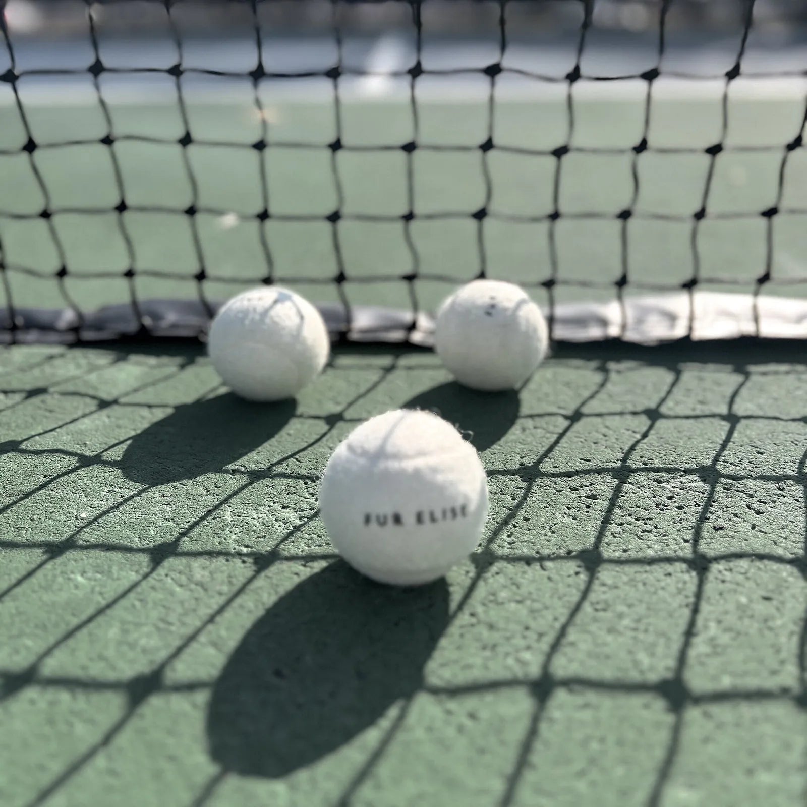 Classic tennis balls in white with "FUR ELISE" text, positioned on a green court under a shadowed net.