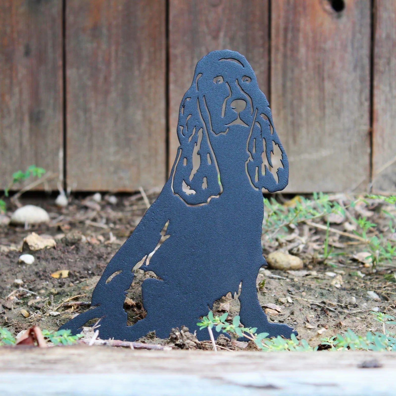 Black Cocker Spaniel metal silhouette sits on the ground, showcasing intricate cutouts, amid greenery and wooden fence bac...