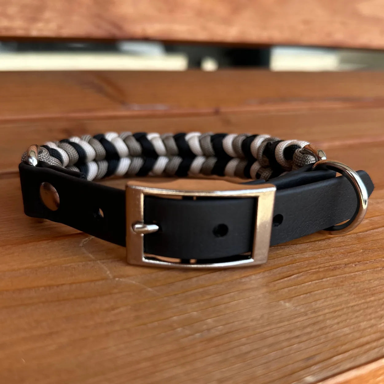 Handmade dog collar in black, tan, and cream with a braided design, nylon paracord, and metal buckle on a wooden surface.