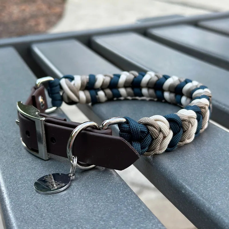 Hand-braided dog collar in navy, tan, and cream with a dark brown strap and silver buckle, placed on a table.