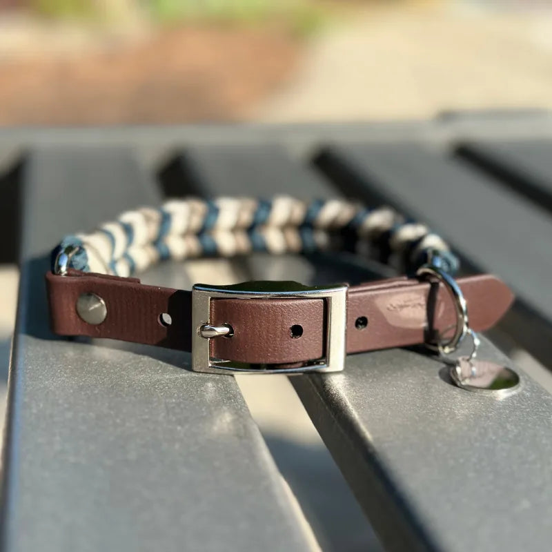 Hand-braided dog collar in navy, tan, and cream, featuring a dark brown strap and silver buckle on a metal table.