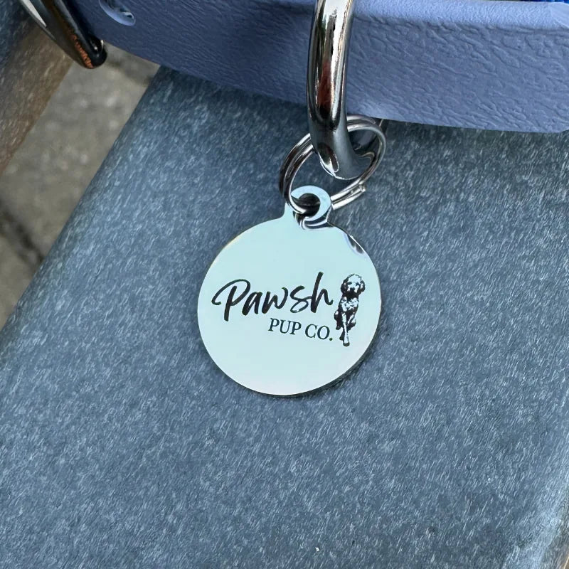 Shiny silver tag with "Pawsh Pup Co." on a royal blue dog collar against a gray surface.