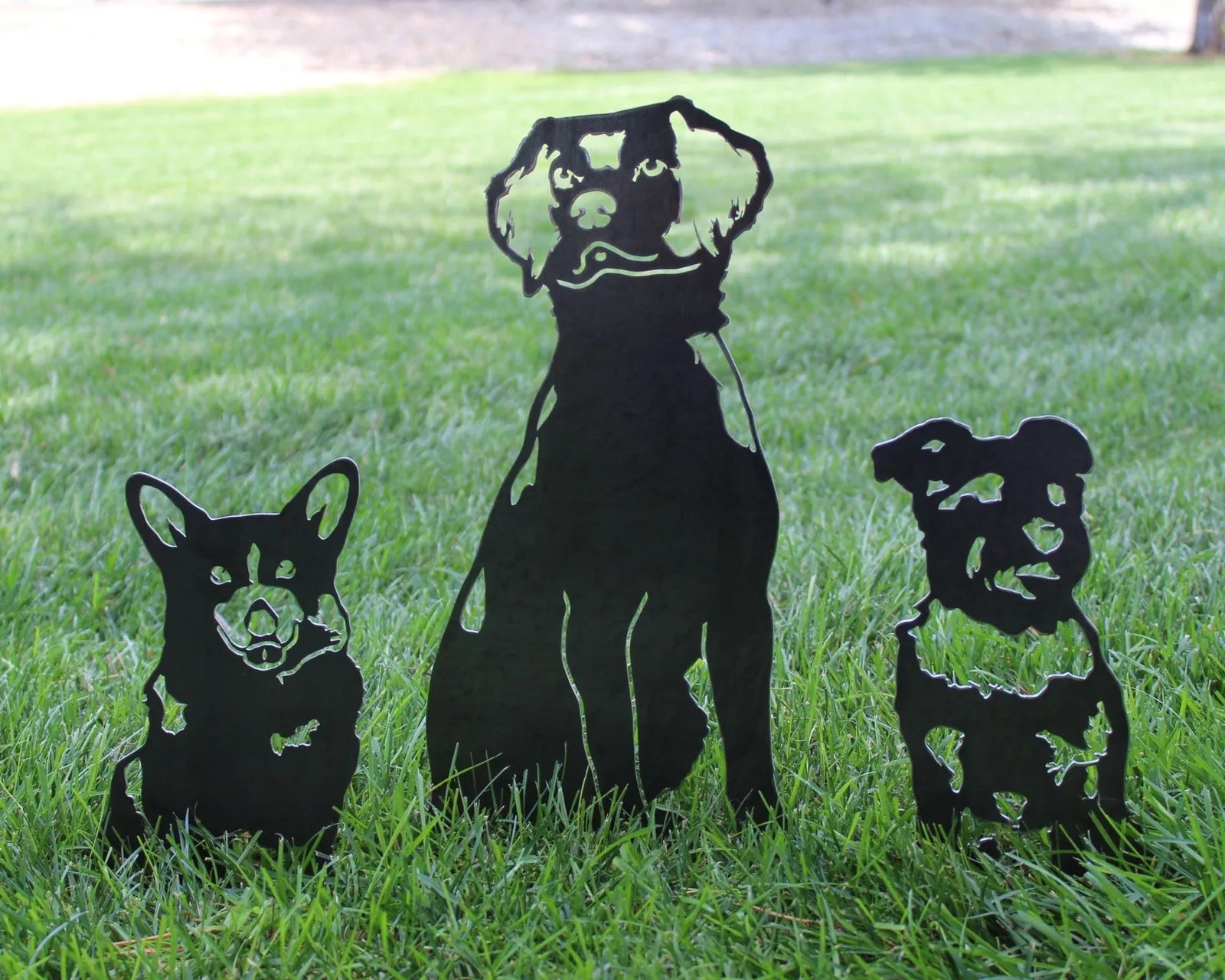 Black metal silhouettes of Corgi, large dog, and smaller dog on green grass, showcasing dog-themed outdoor art.
