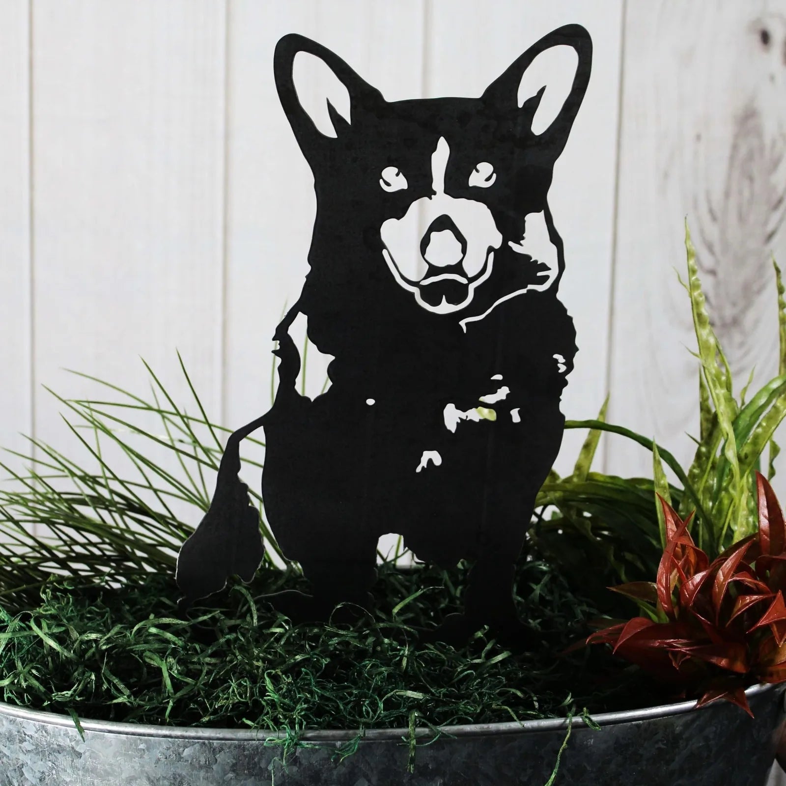 Corgi silhouette in matte black metal, showcasing detailed features, surrounded by greenery in a garden setting.
