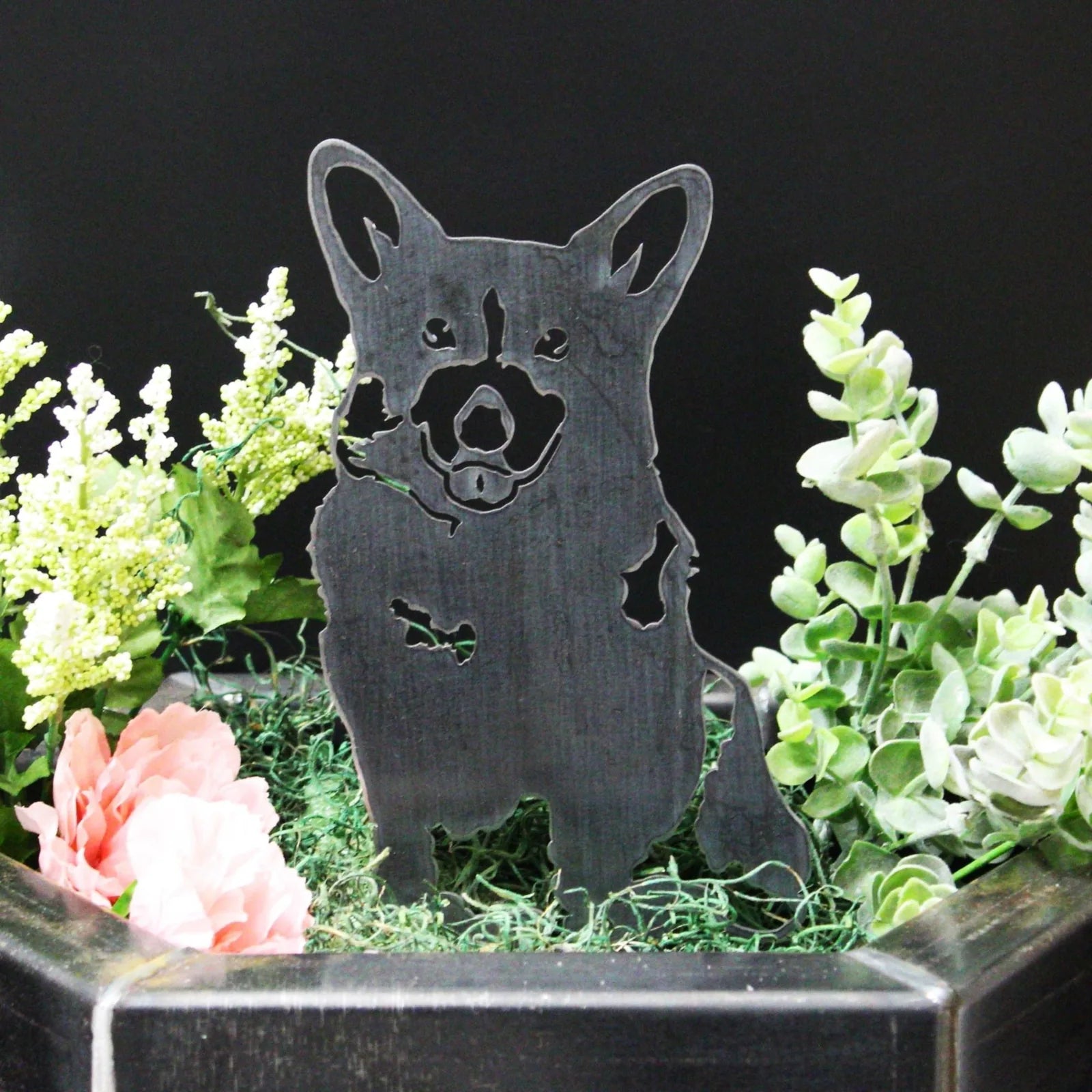Black metal Corgi silhouette art nestled among green foliage and pink flowers in a planter. Ideal outdoor decor.