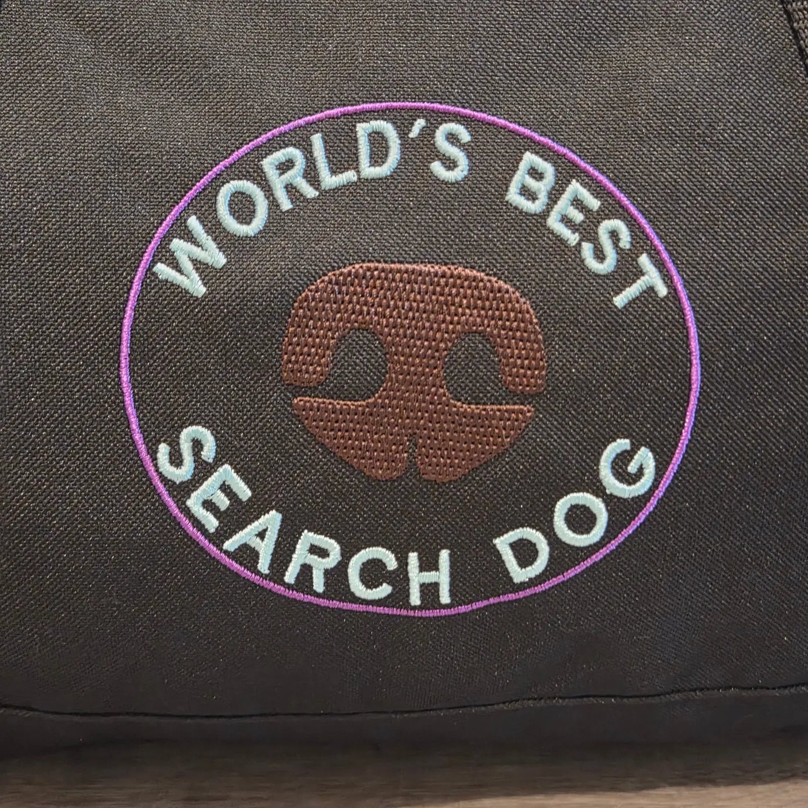 Custom embroidered "World's Best Search Dog" logo on a black duffel bag, featuring a nose silhouette in brown and light blue.